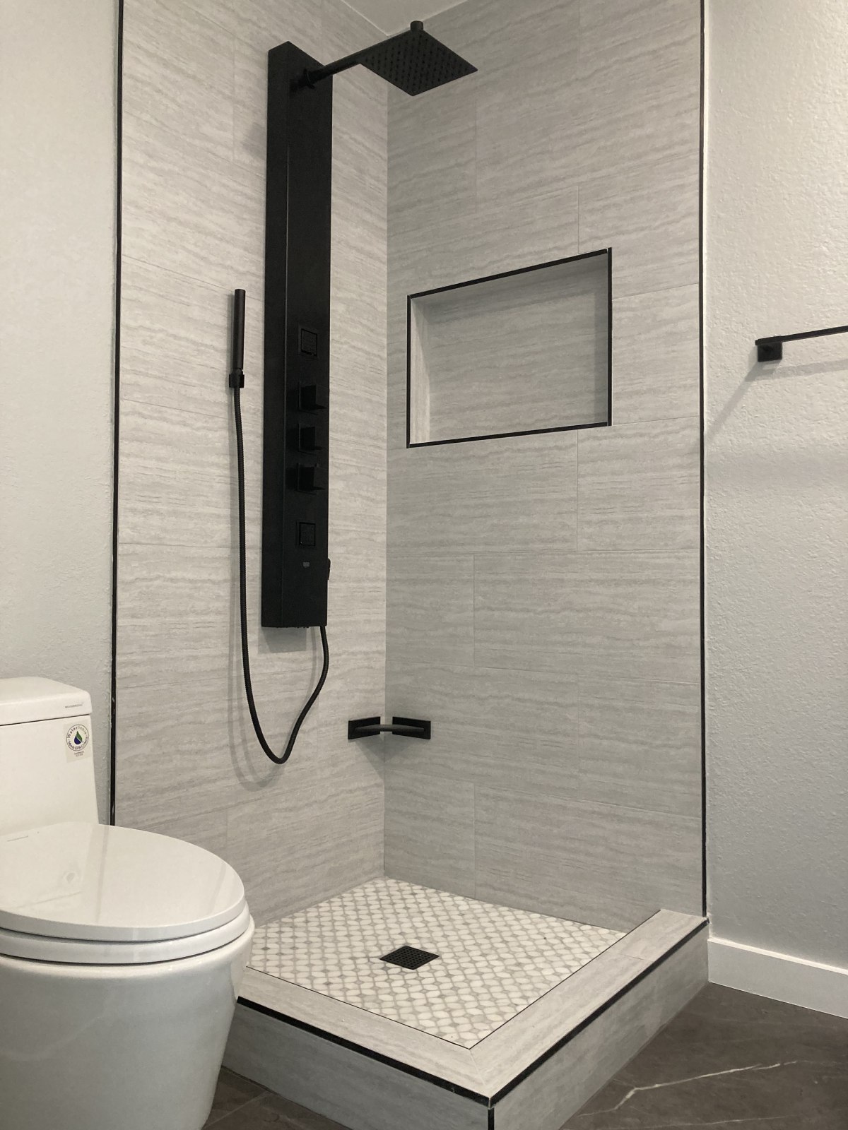 Modern bathroom with gray textured tile walls, black rainfall shower system, white mosaic tile shower floor, and white toilet