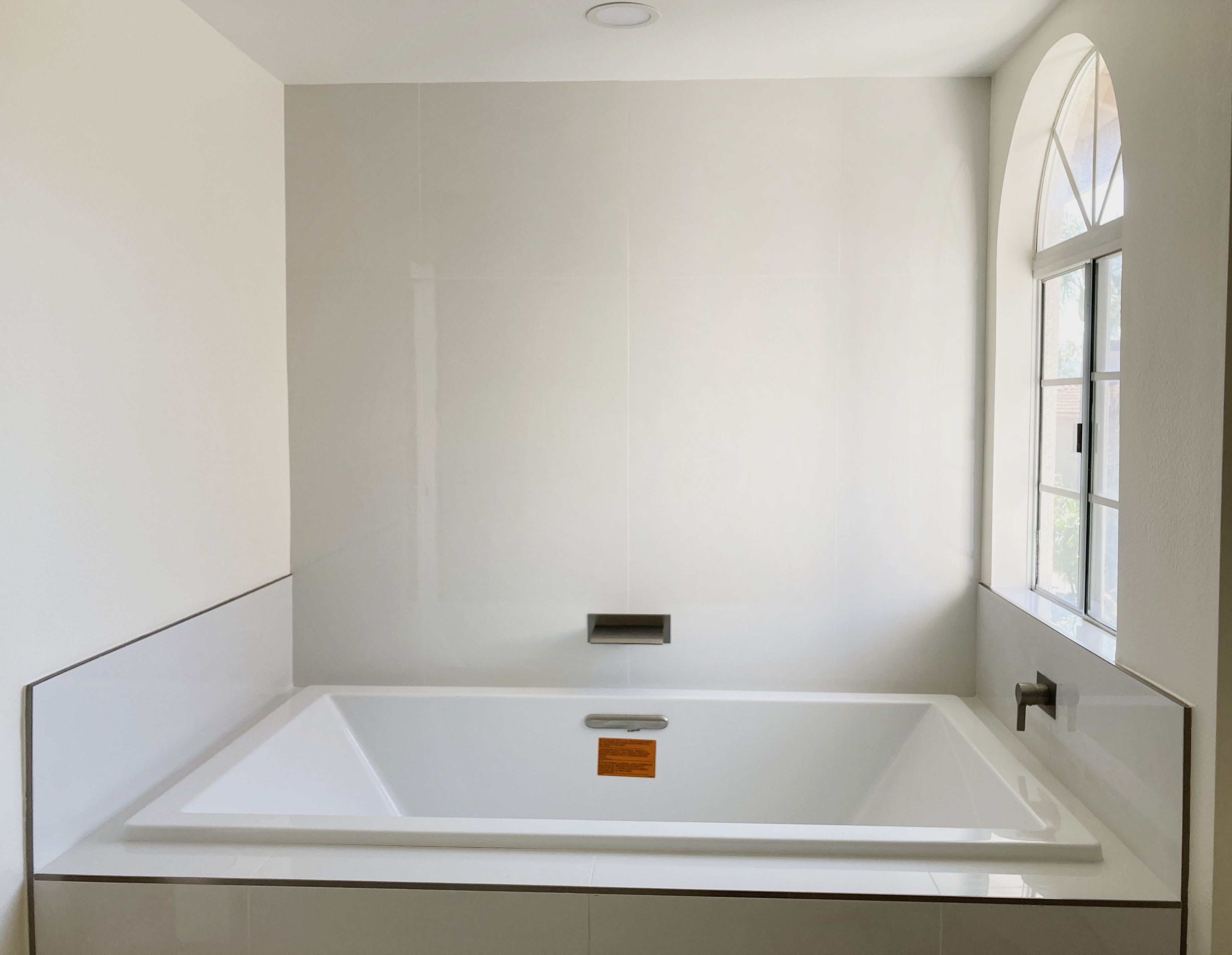Modern white rectangular bathtub in minimalist bathroom with arched window and neutral gray walls