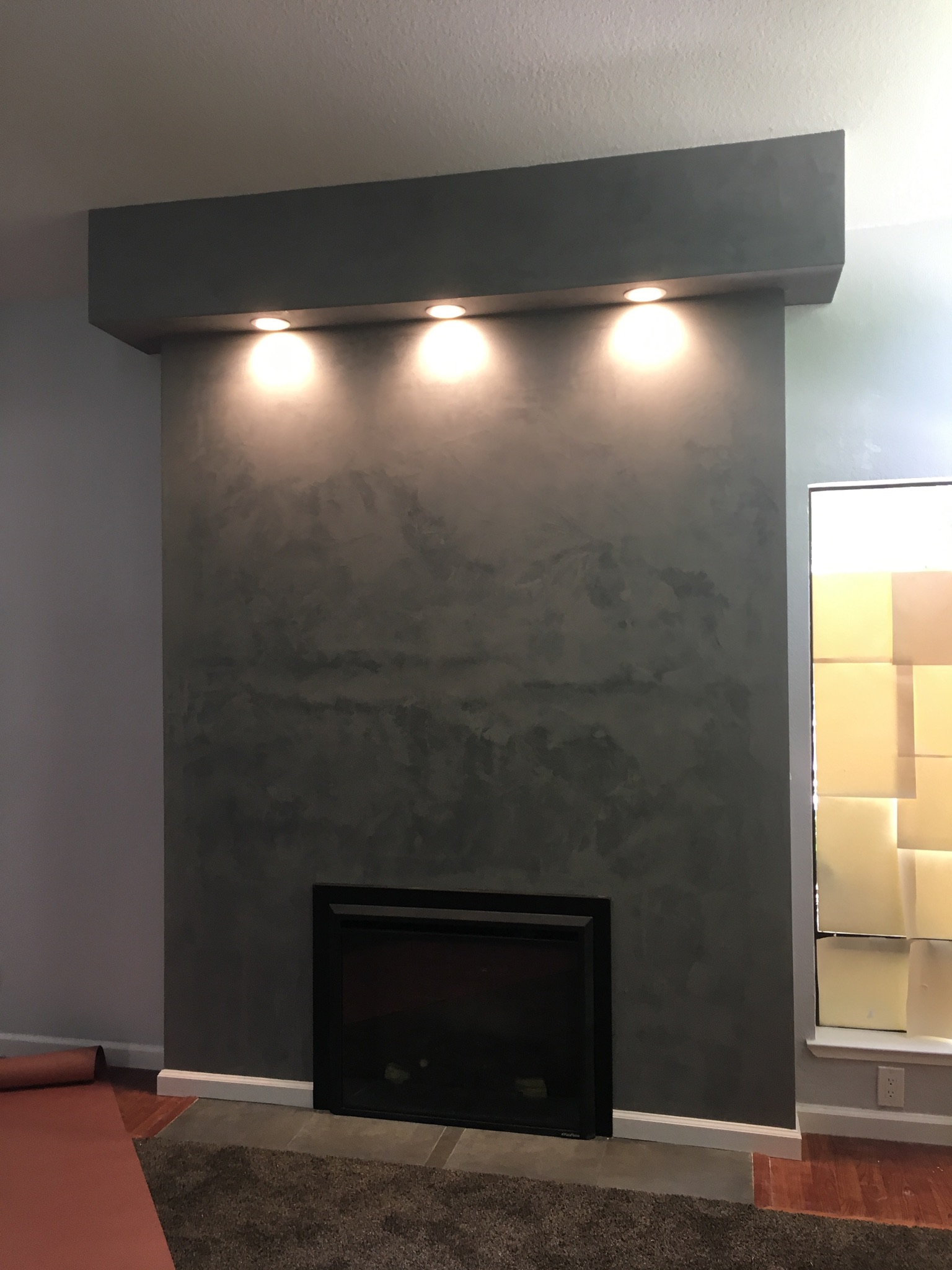 Modern concrete fireplace with three recessed LED spotlights in dark gray mantel, black firebox opening, and textured stone surround