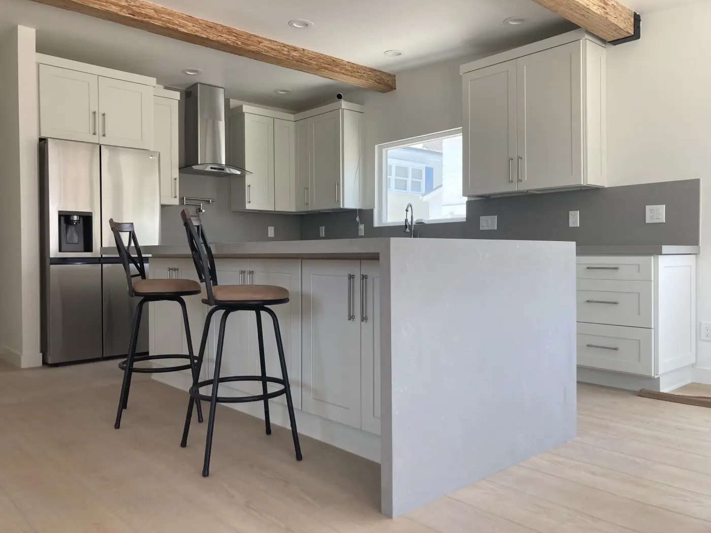 Modern white kitchen with gray backsplash, wood beam ceiling, stainless appliances, and black bar stools at island