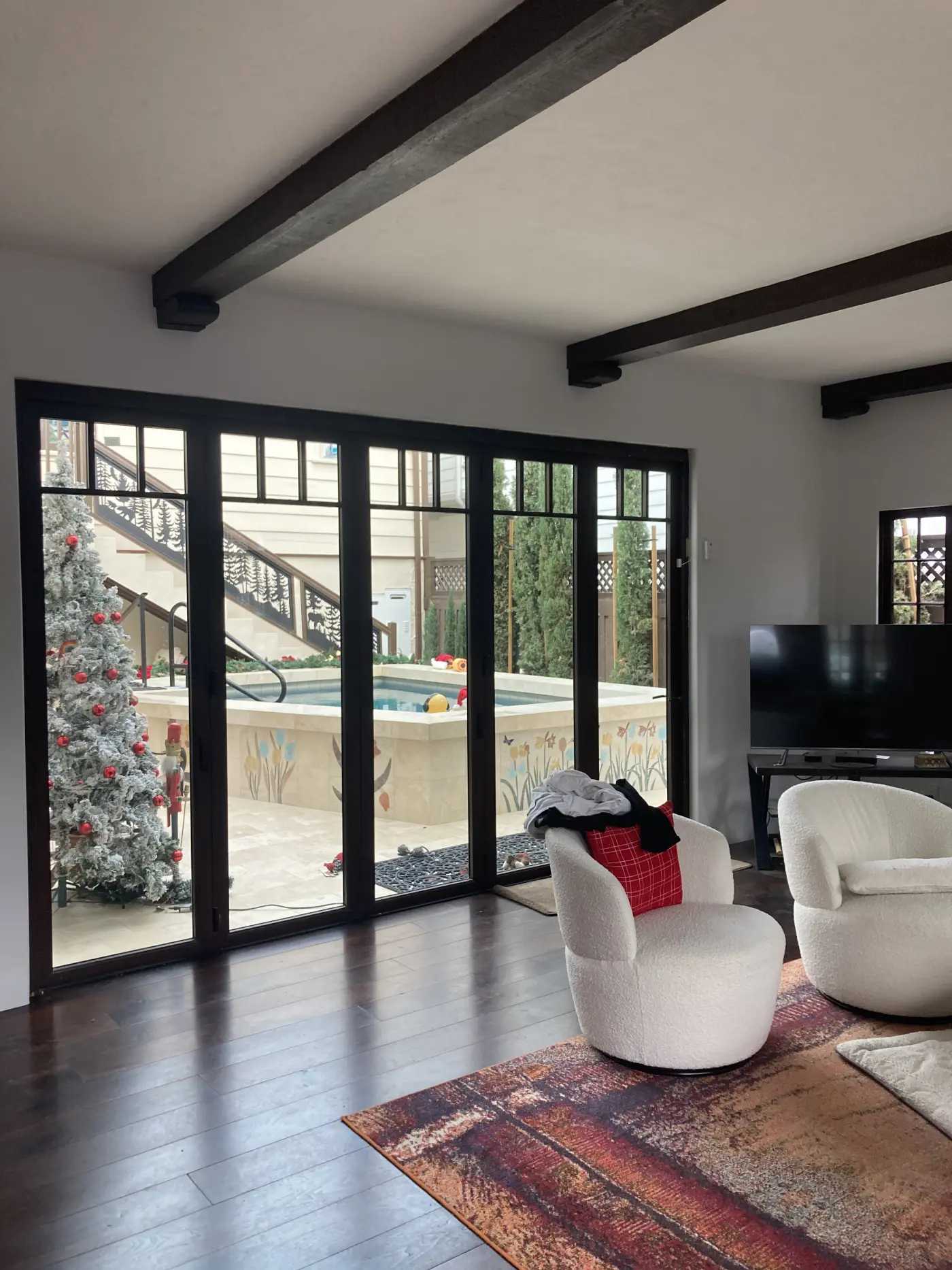Modern living room with exposed dark ceiling beams, large black-framed windows, white swivel chairs, and view of outdoor pool area