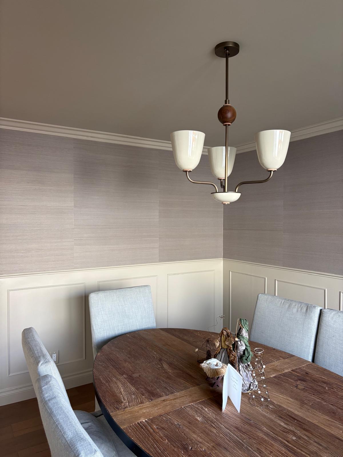 Modern dining room with round wood table, gray upholstered chairs, white wainscoting, gray grasscloth wallpaper, and mid-century chandelier