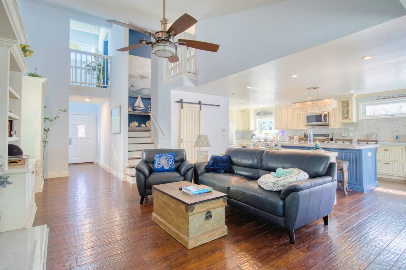 Open-concept living room with dark leather sofa, hardwood floors, white kitchen, nautical decor, and exposed ceiling beams
