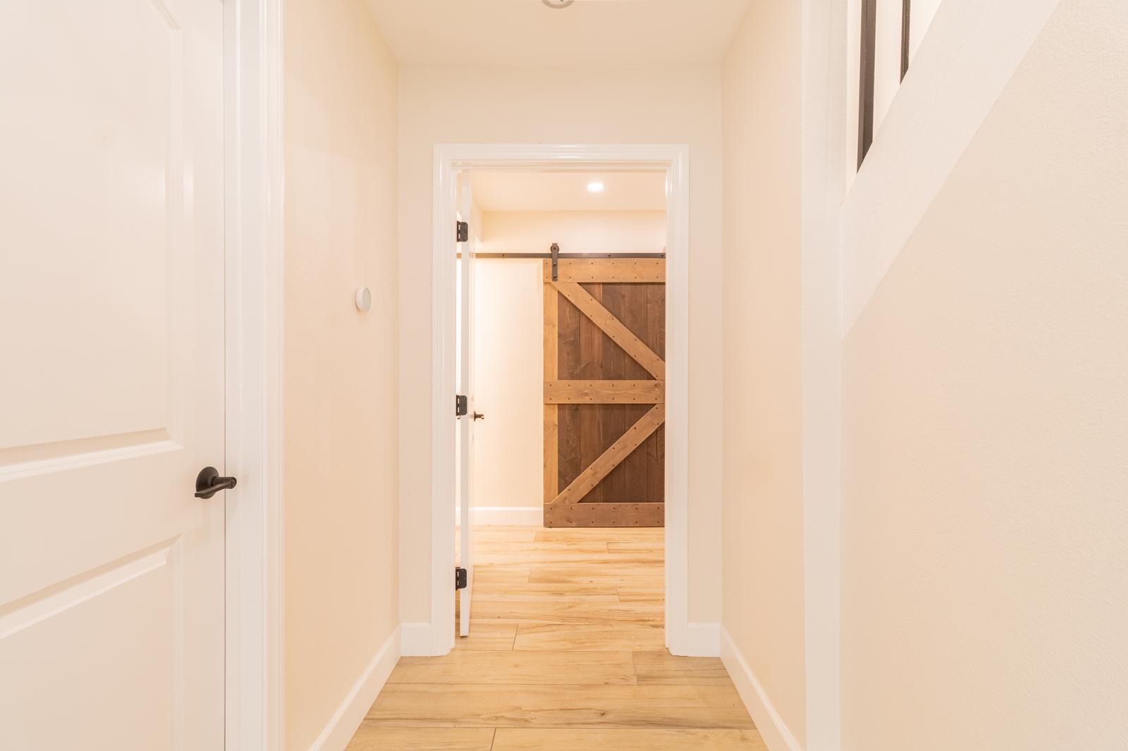 Clean white hallway with light wood floors leading to rustic barn door with diagonal wood planking and black hardware