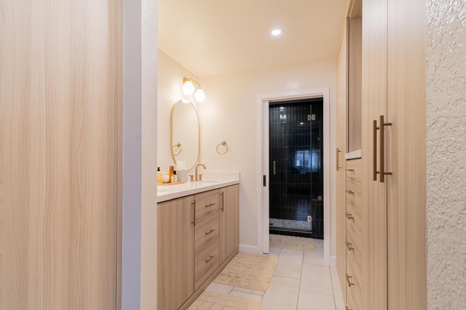 Modern bathroom with light wood vanity, oval mirror, brass fixtures, and dark tile shower with glass door