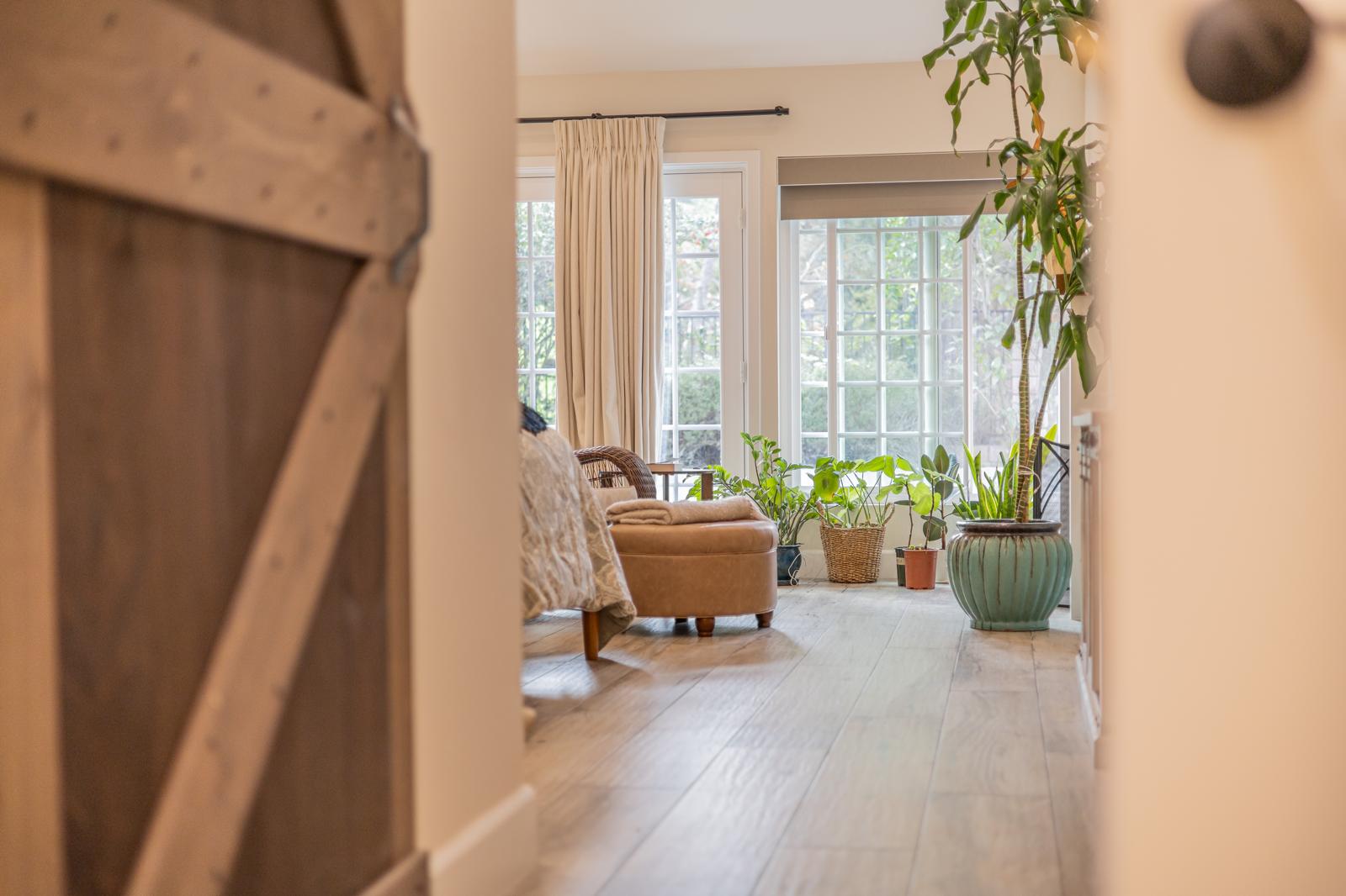 Bright modern living room with exposed wood beams, light hardwood floors, beige curtains, wicker chair, and potted plants by large windows