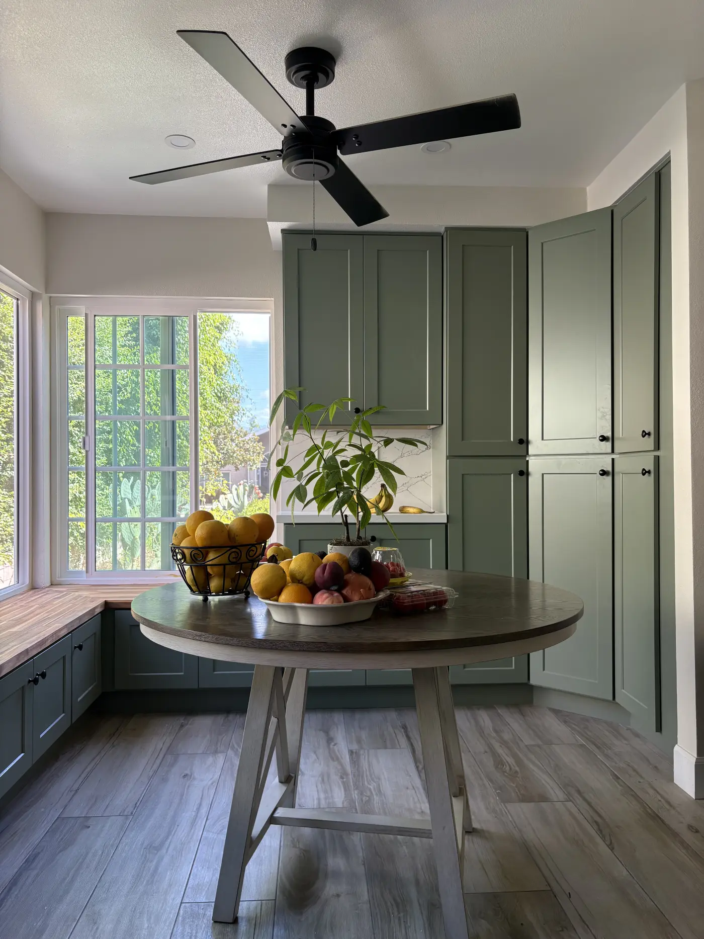 Modern kitchen with sage green cabinets, round wooden dining table with fresh fruit, black ceiling fan, and large windows overlooking greenery