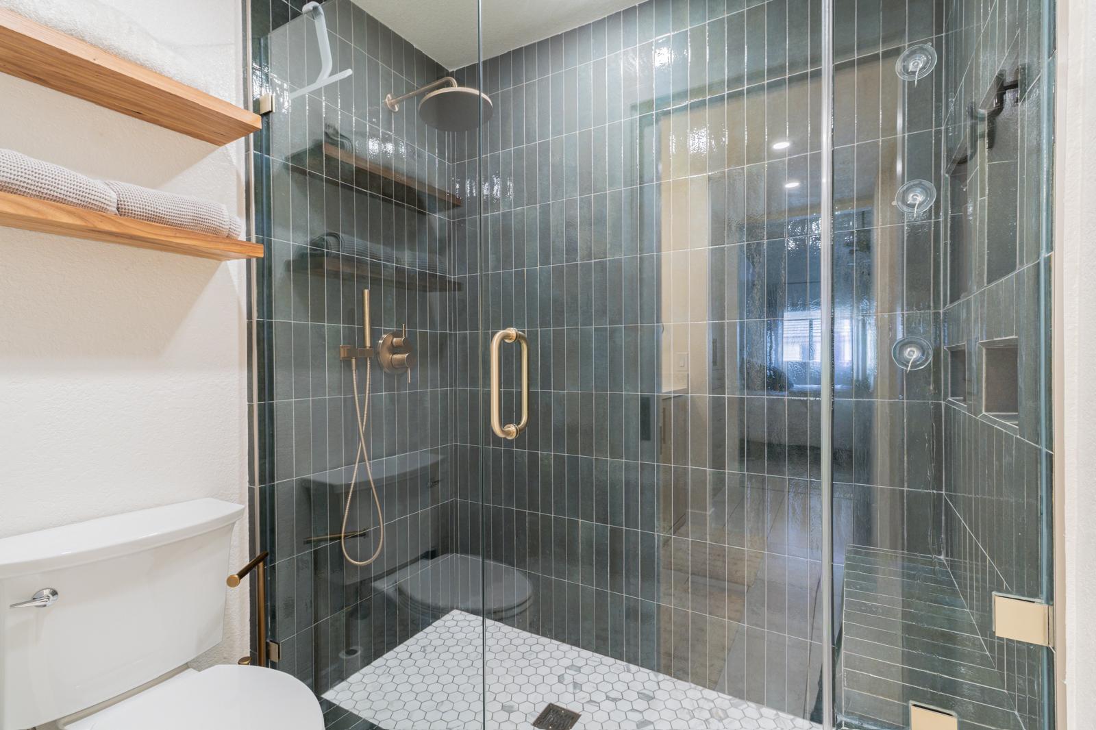 Modern bathroom with green vertical tile shower, brass fixtures, glass doors, white hexagon floor, and floating wood shelves