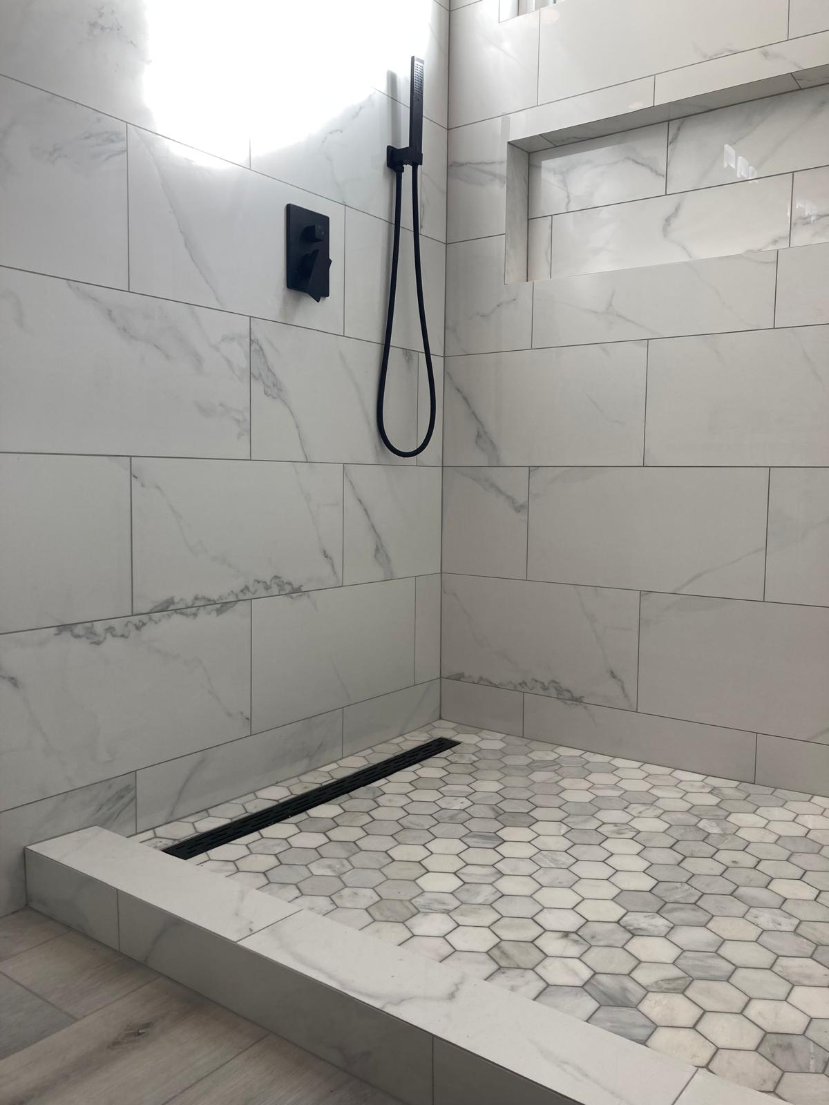 Modern walk-in shower with white marble-look tile walls, hexagonal mosaic floor, black linear drain, and matte black handheld shower fixture