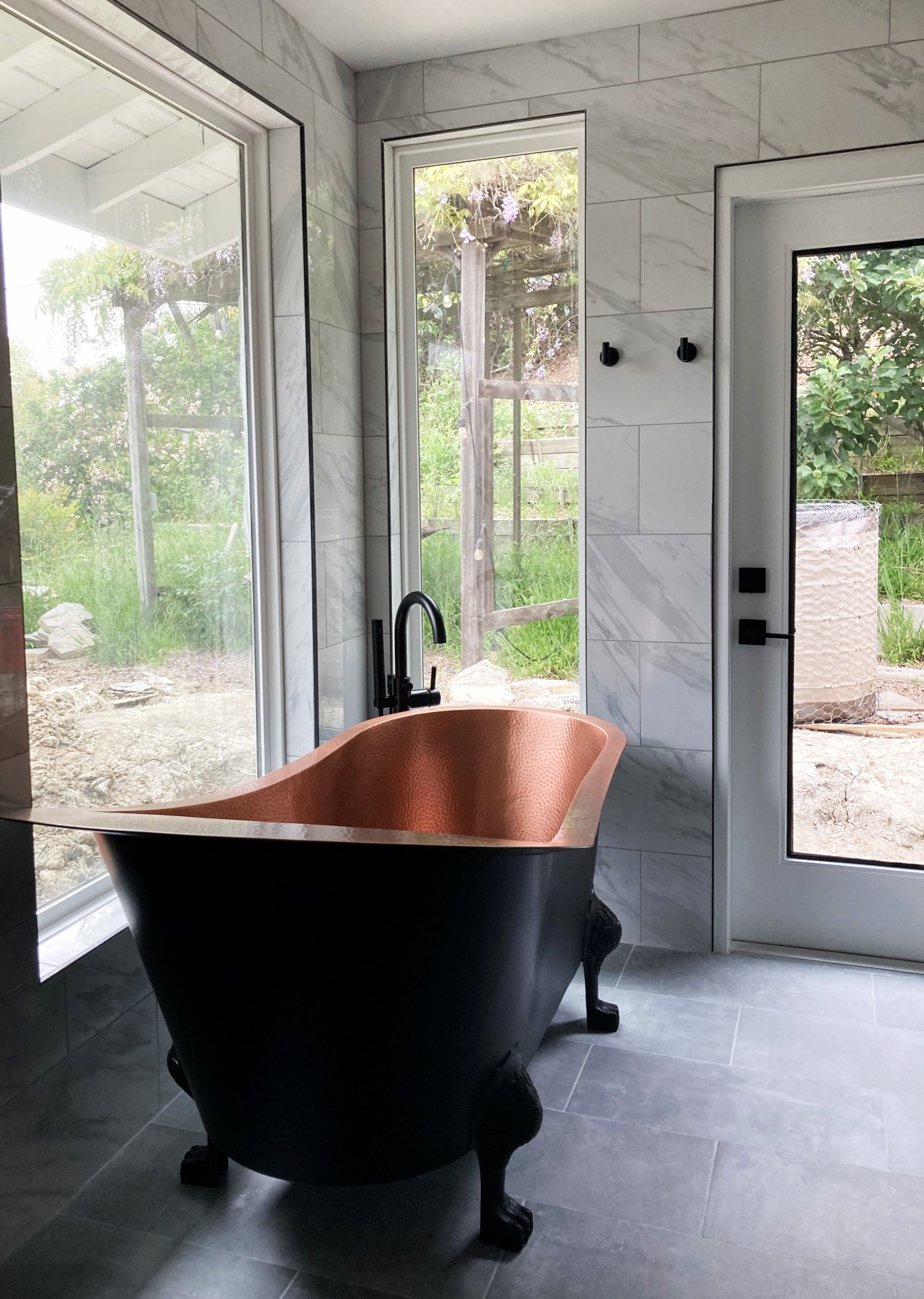 Modern bathroom with copper interior clawfoot tub, black exterior, marble wall tiles, and large windows overlooking garden