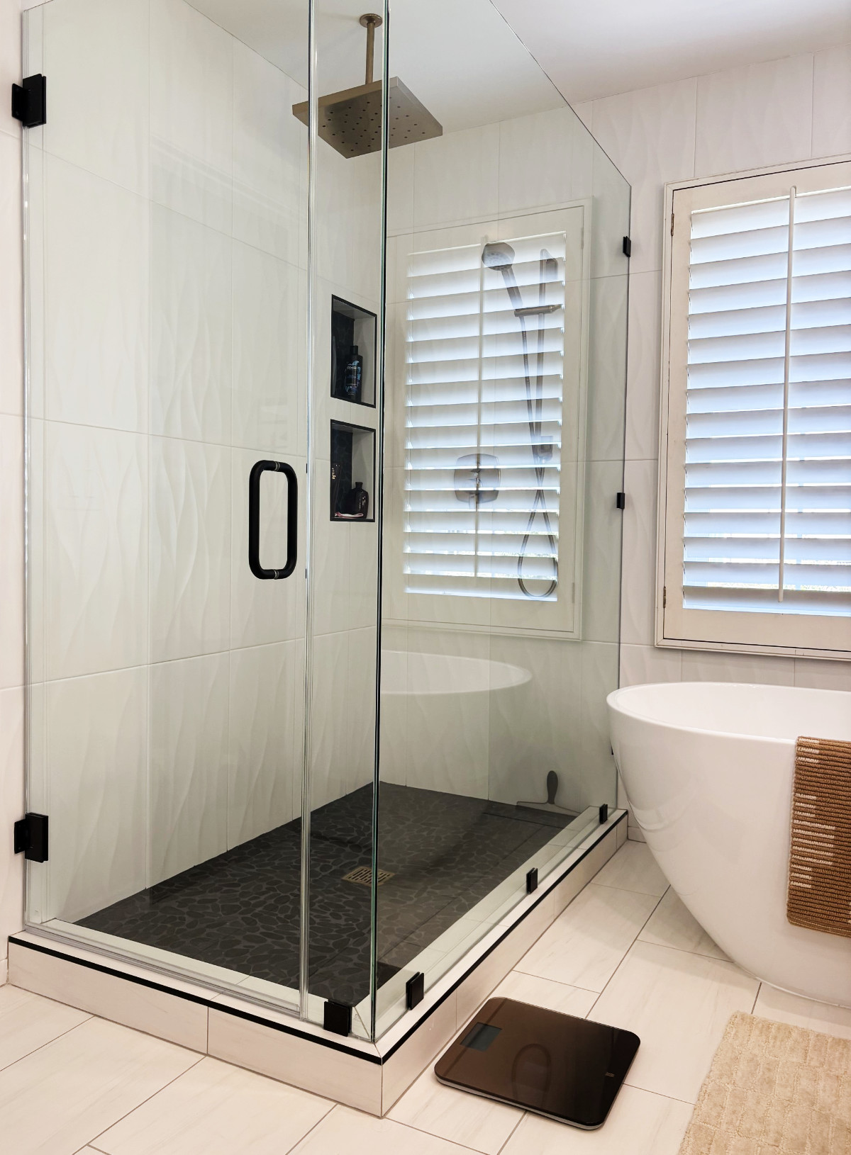 Modern bathroom with glass-enclosed shower, dark stone floor, white freestanding tub, and plantation shutters