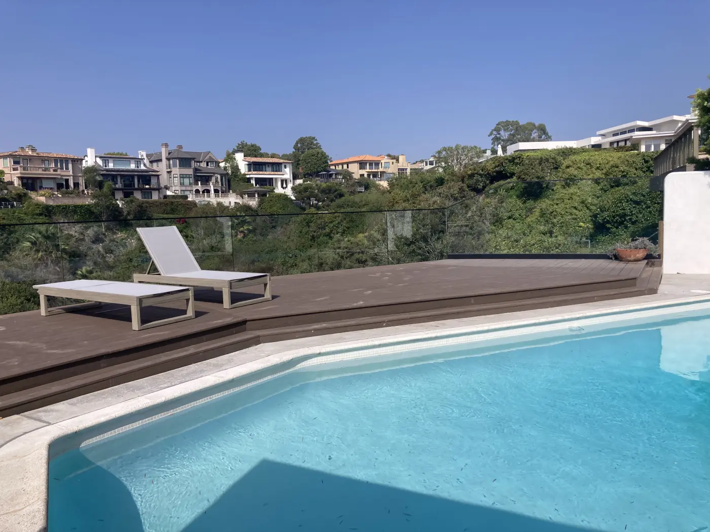 Modern hillside pool deck with white lounge chairs, glass railing, and luxury homes overlooking canyon landscape