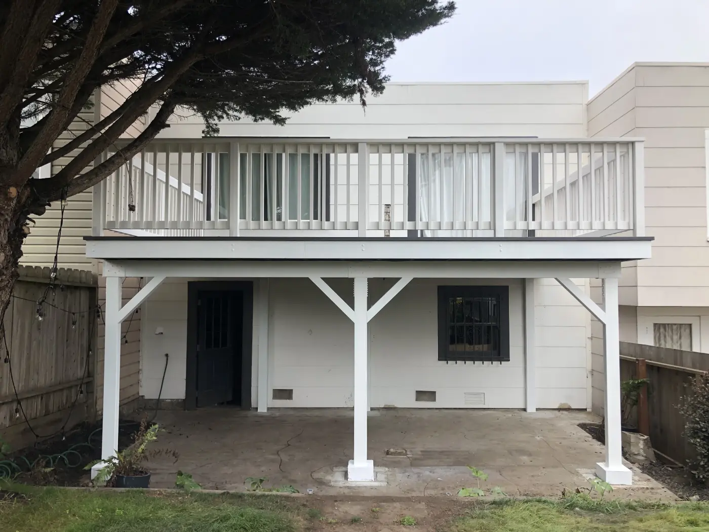 White wooden deck with railing above covered patio area, supported by white posts, attached to beige siding home