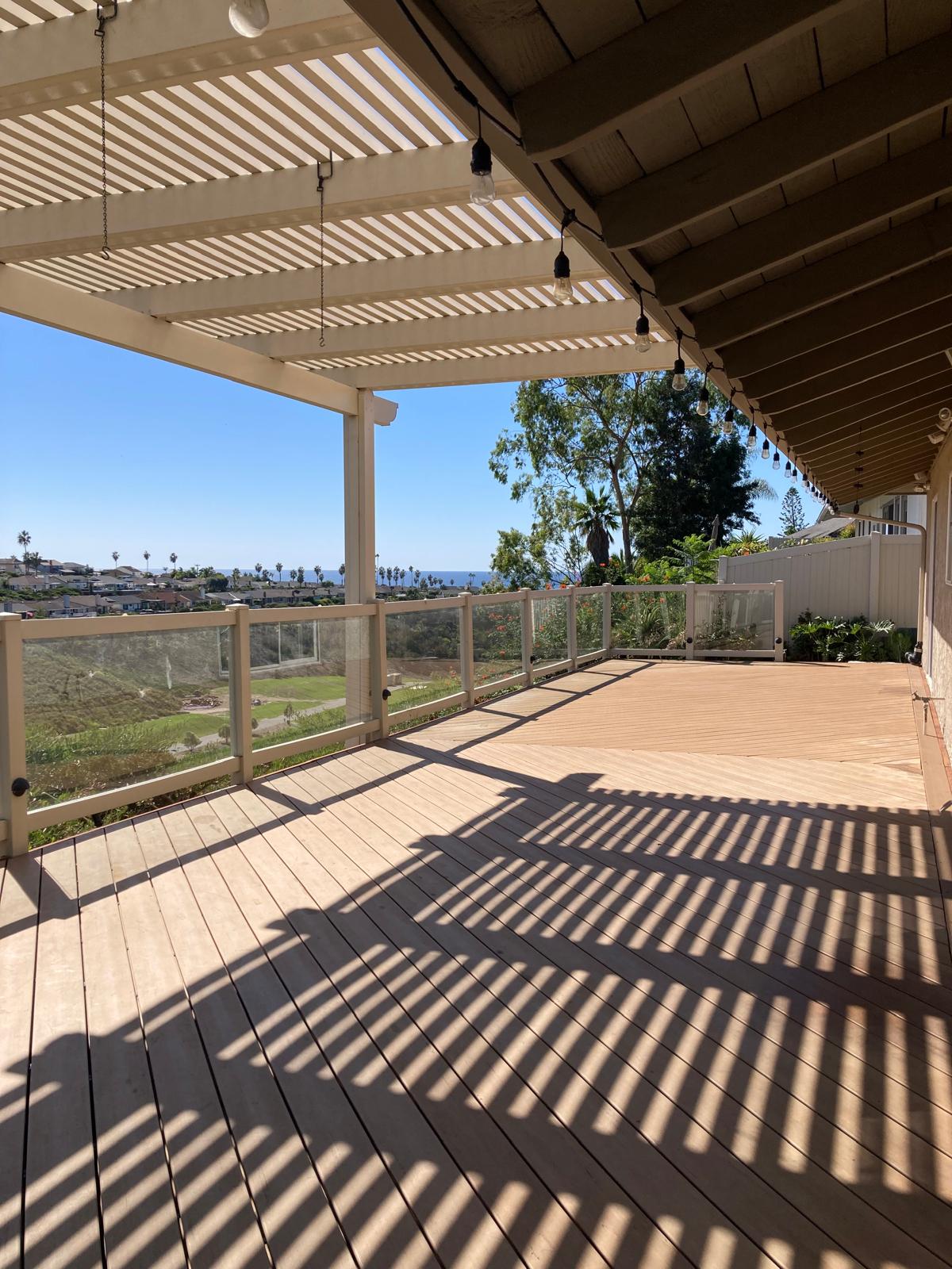 Spacious wooden deck with white pergola casting striped shadows, glass railings, pendant lighting, and ocean view in distance