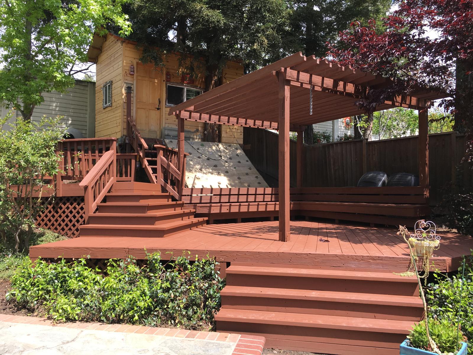 Multi-level redwood deck with pergola, built-in slide, stairs, and railings surrounded by mature trees and landscaping