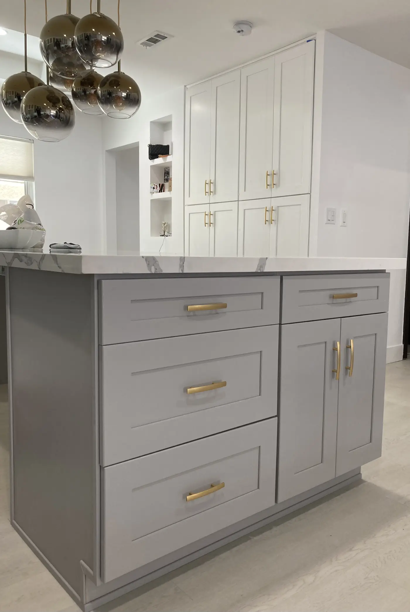 Modern kitchen island with gray shaker cabinets, brass hardware, white quartz countertops, and smoky glass pendant lights