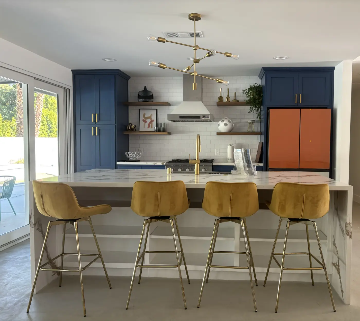 Modern kitchen remodel with navy blue cabinets, orange accent panels, white marble island, gold bar stools, and brass lighting fixture