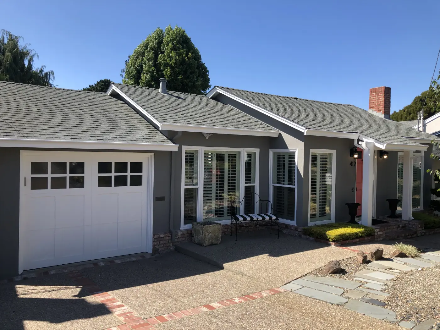 Single-story gray ranch home with white garage door, plantation shutters, brick chimney, and drought-tolerant landscaping
