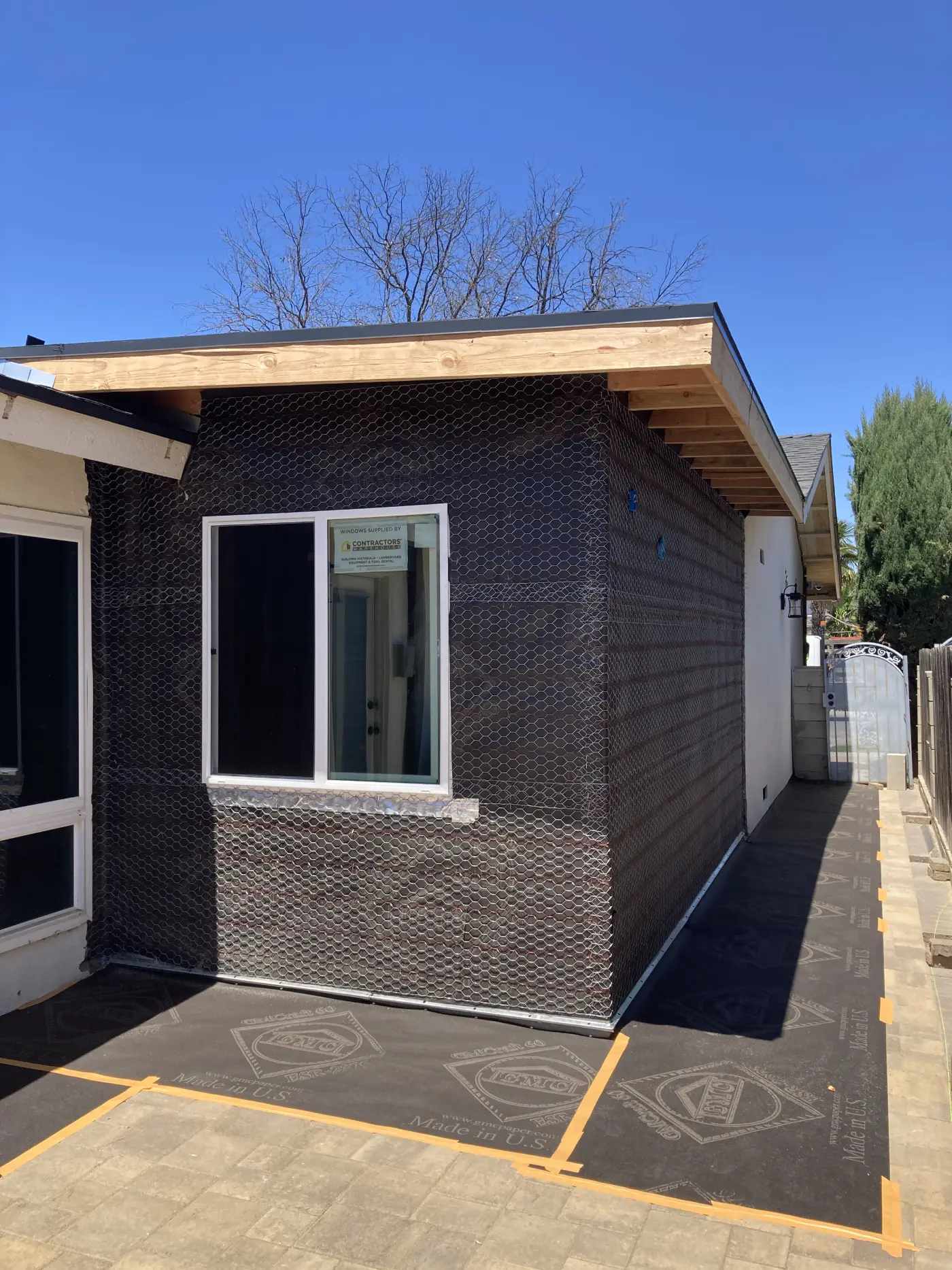 Modern home addition under construction with black metal mesh siding, exposed wood beams, white window frame, and clear blue sky