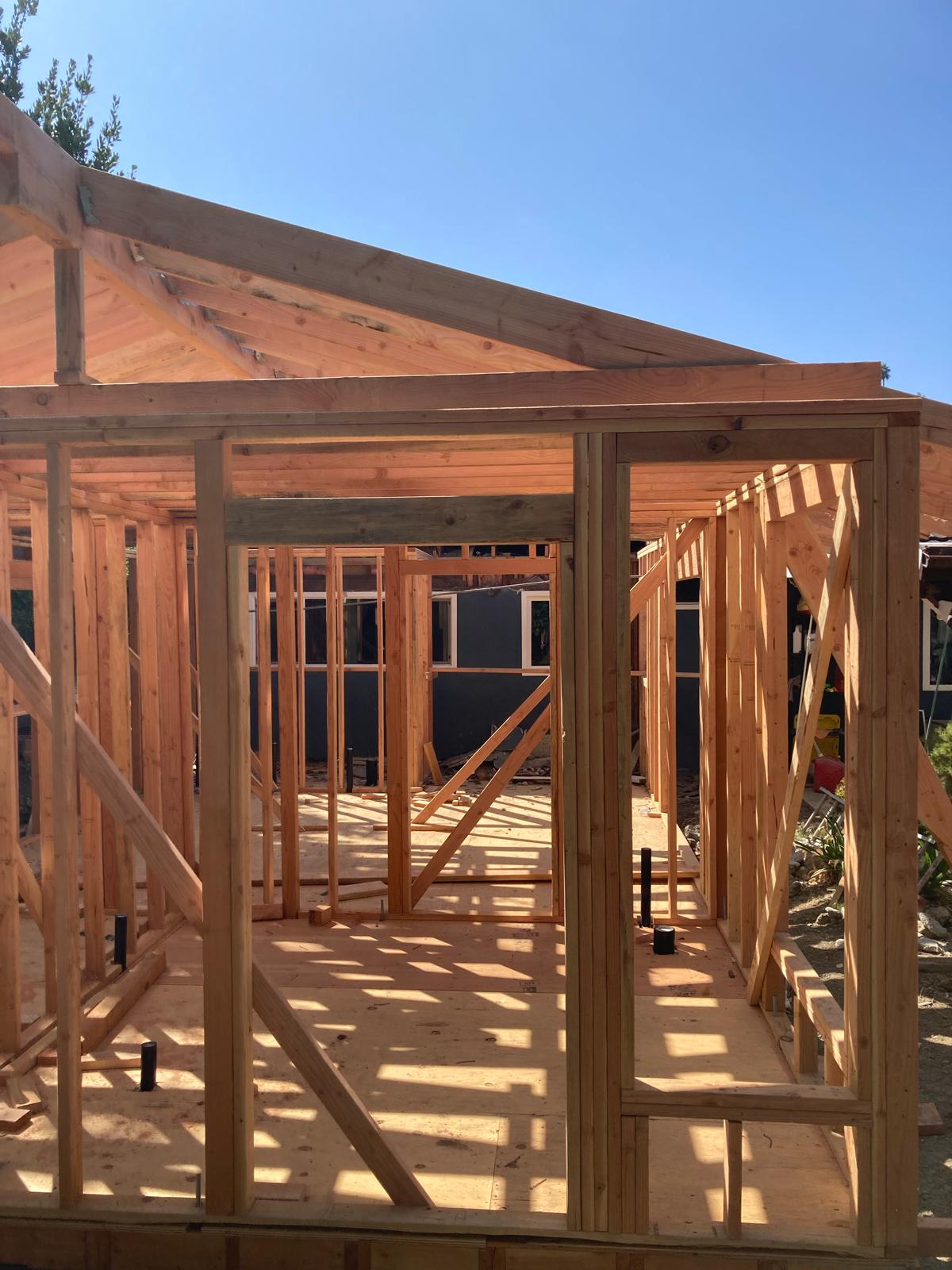 Wood frame construction site showing natural lumber framing, roof trusses, and window openings under clear blue sky
