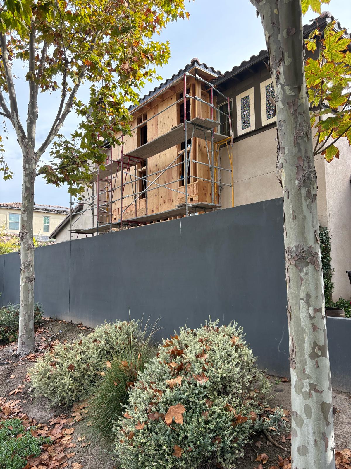 House under construction with exposed wood framing, scaffolding, and stucco exterior behind gray privacy fence in residential neighborhood