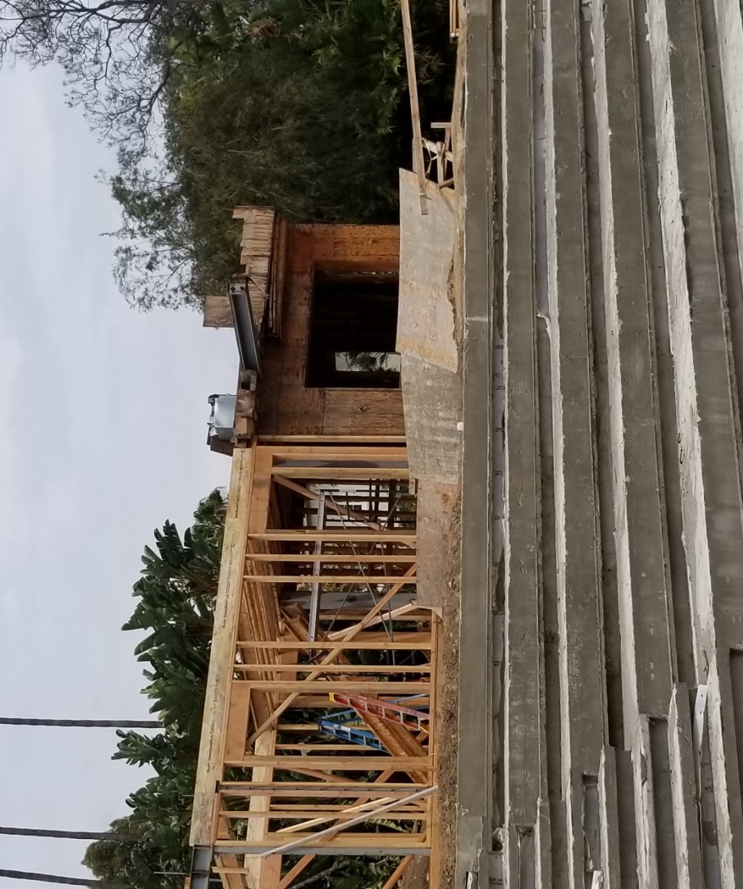 Wood-framed house under construction with exposed studs and concrete foundation, ladders and construction equipment visible