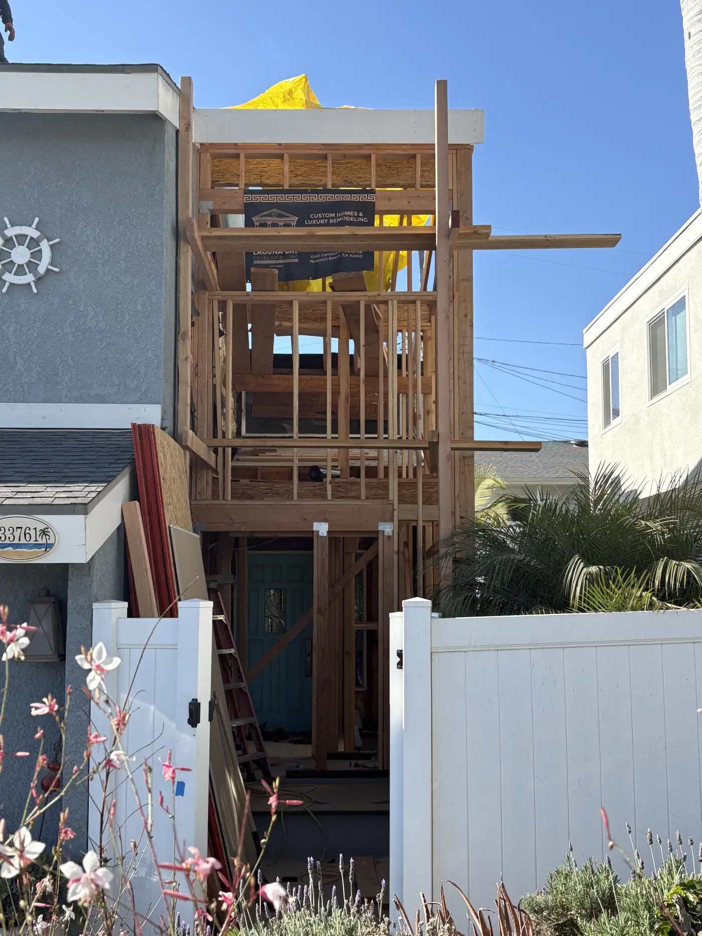 Multi-story home under construction with exposed wooden framing, yellow construction material on roof, white fence in foreground