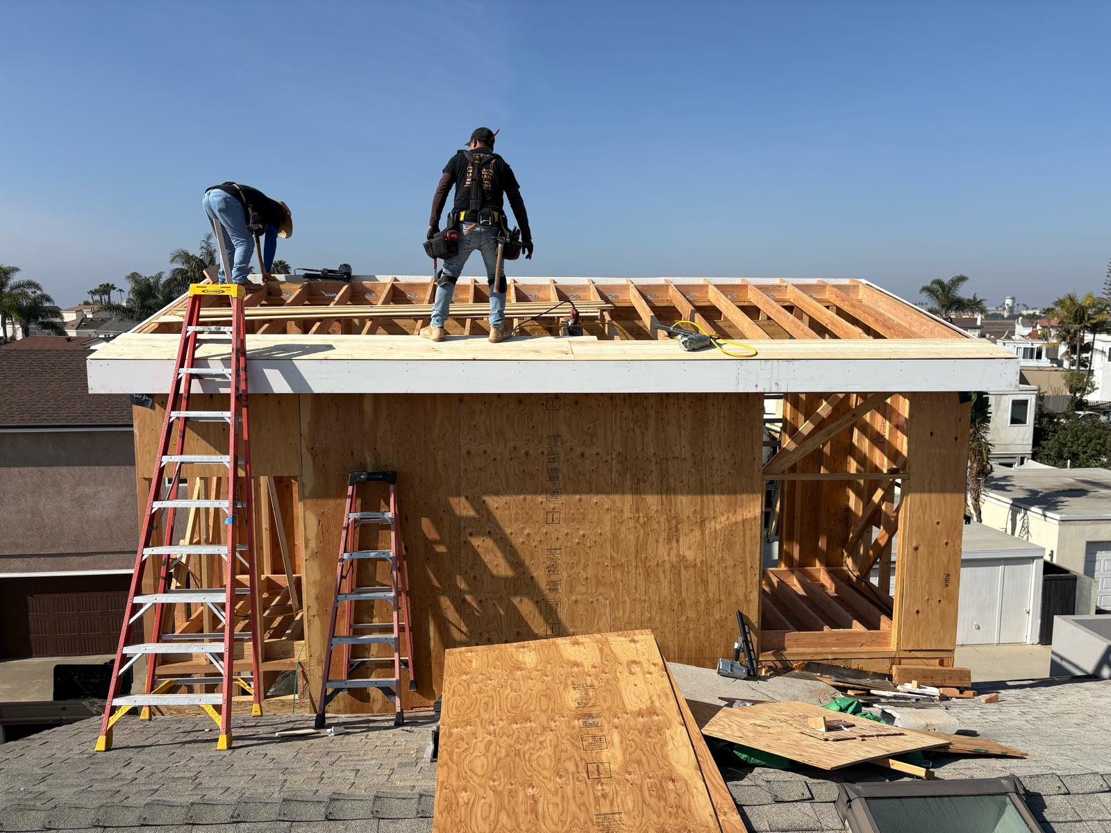 Two construction workers building wooden frame structure on rooftop with exposed rafters, plywood sheathing, and red ladders