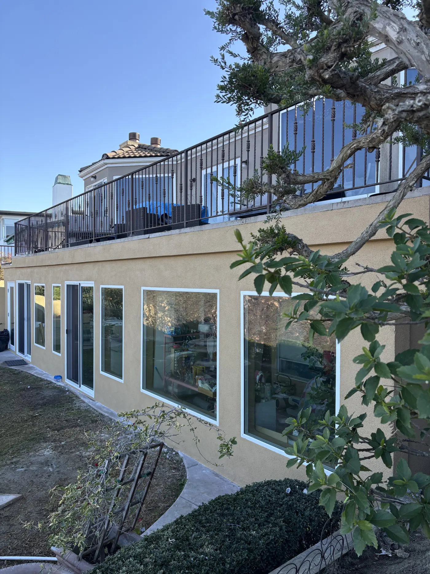 Two-story beige stucco home with white-framed windows, black metal balcony railing, and mature tree in landscaped yard
