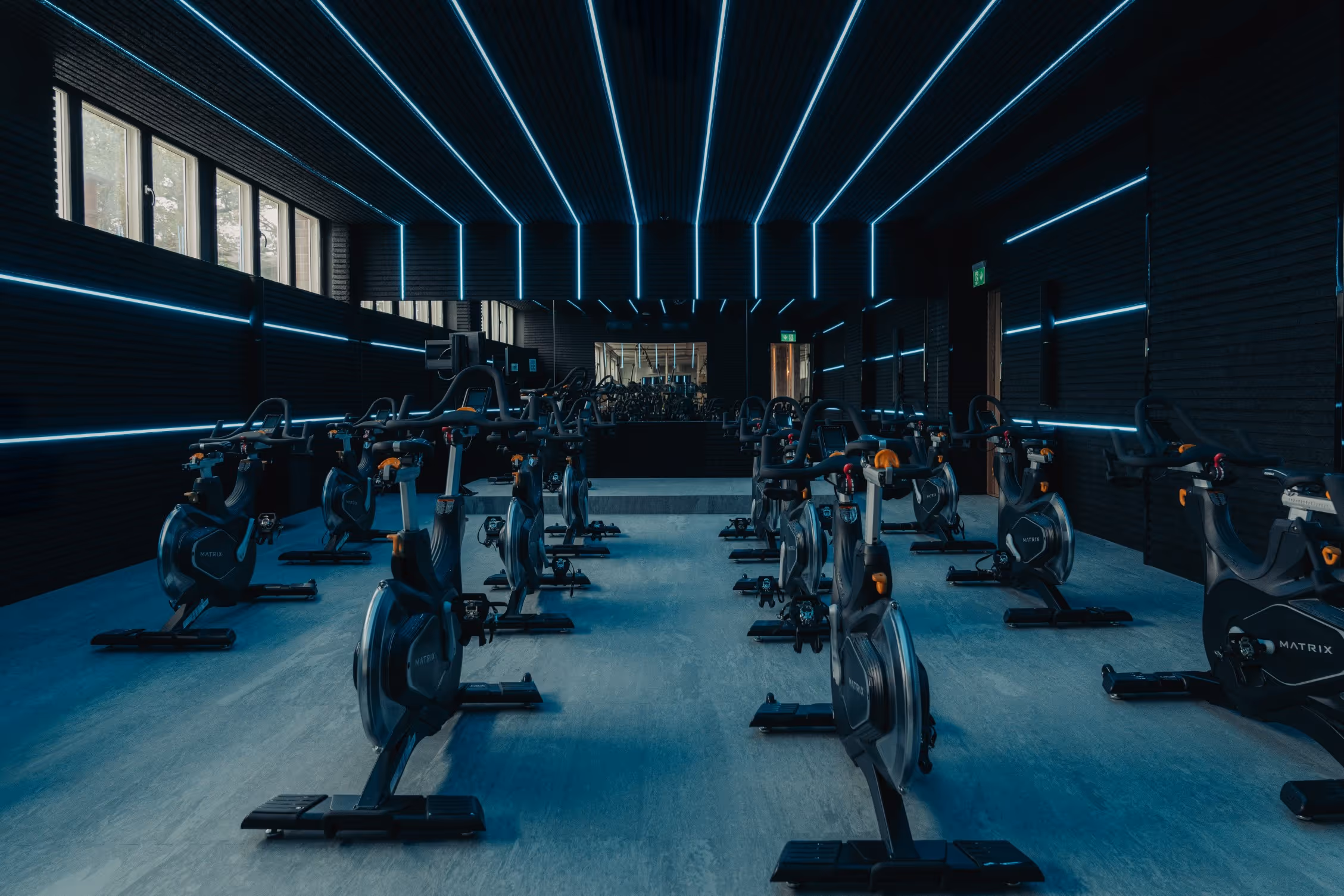 Indoor cycling studio with stationary bikes arranged in rows under blue LED strip lights on the ceiling and walls.