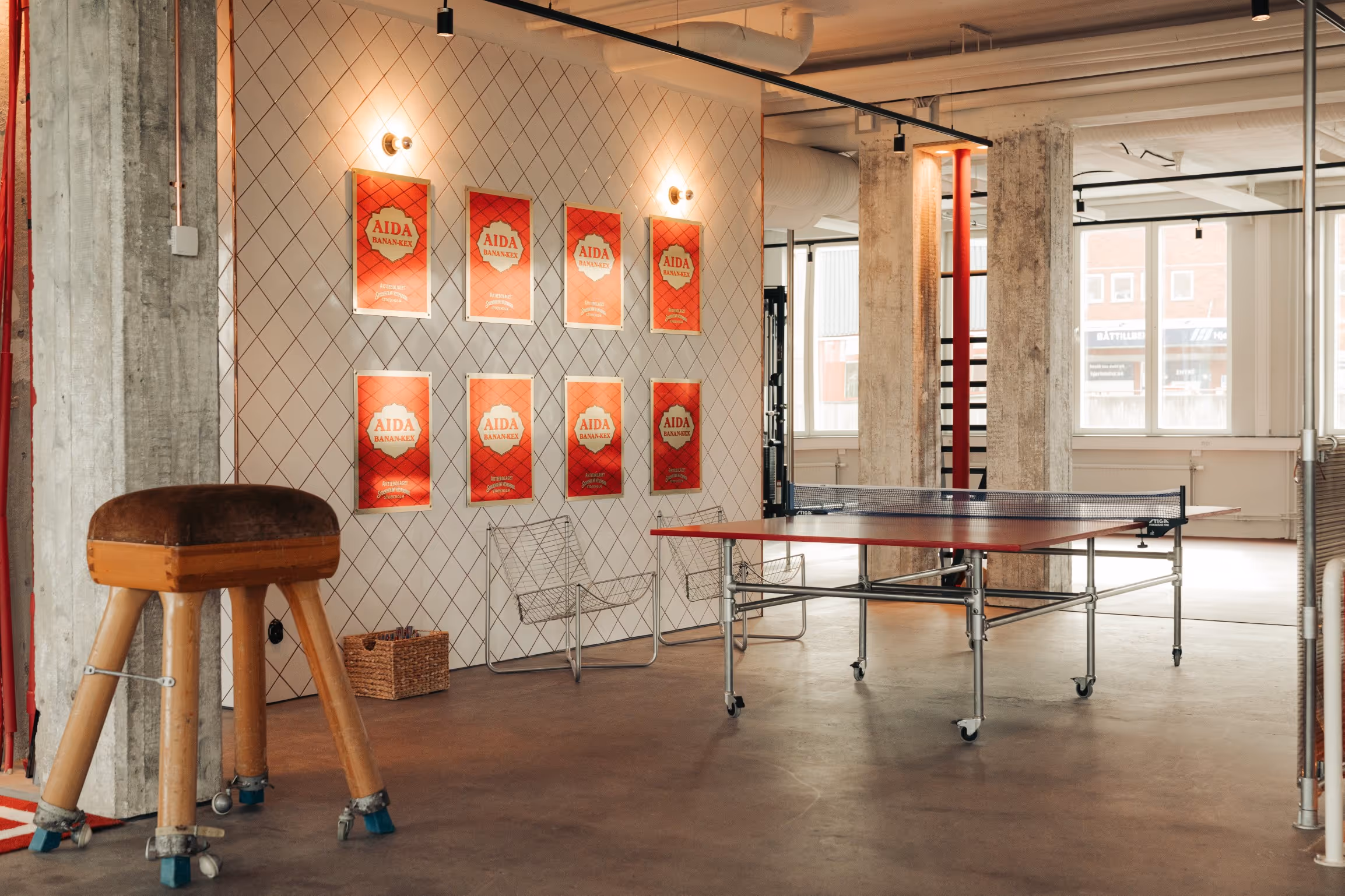 Modern indoor recreation space with a ping pong table, vintage wooden vaulting horse, wire chairs, and red AIDA Banan-Kex posters on a tiled wall.