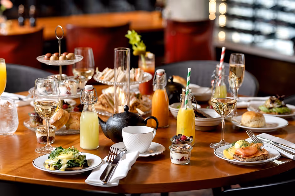 Table set with elegant brunch featuring eggs Benedict with smoked salmon, green vegetable dish with hollandaise, assorted sandwiches, pastries, parfaits with berries, various juices, wine glasses, and tea pot.