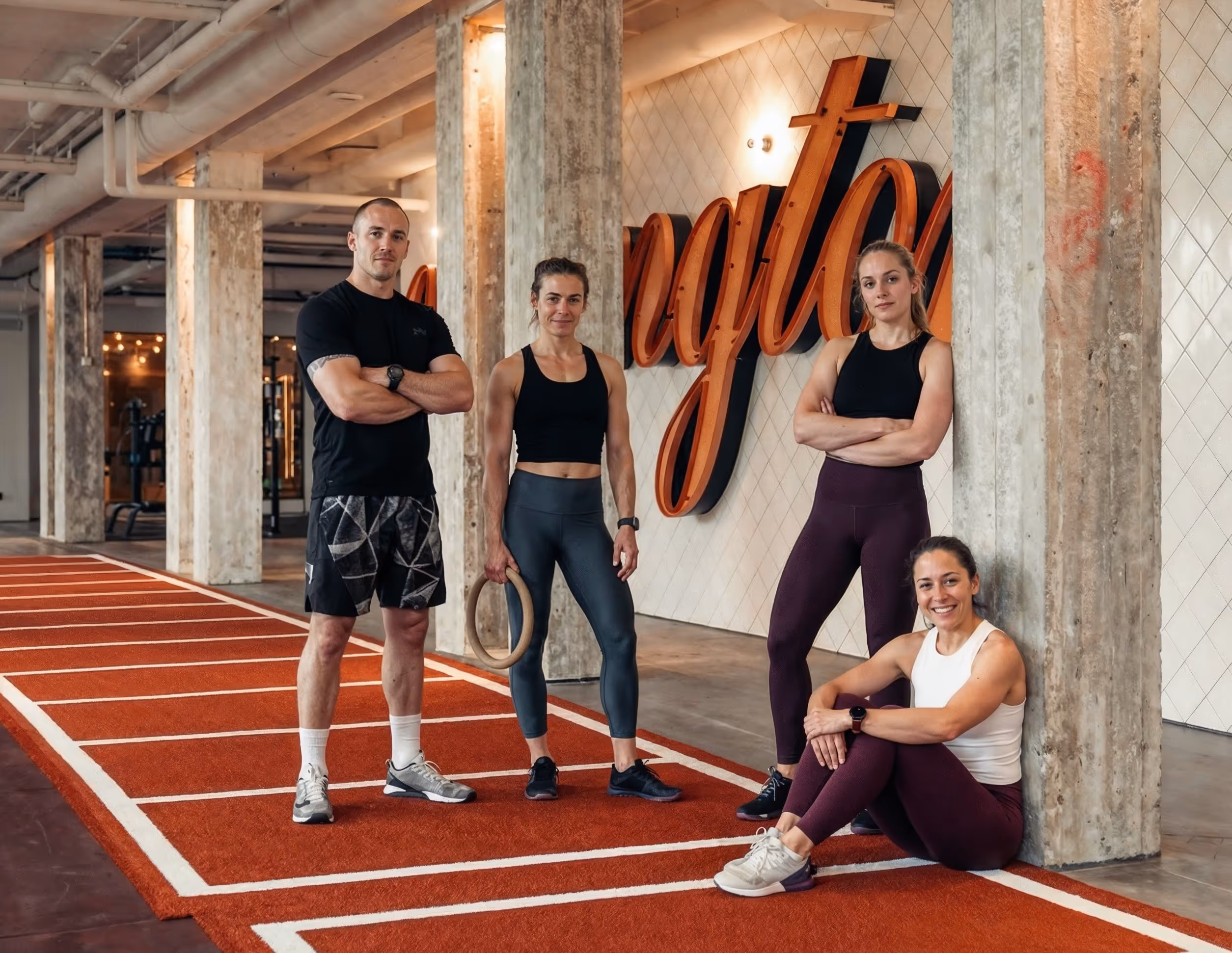 Group of four fit adults posing in a modern gym with concrete pillars and a red running track, two women standing with arms crossed, one woman holding a gymnastic ring, and another woman sitting against a pillar.