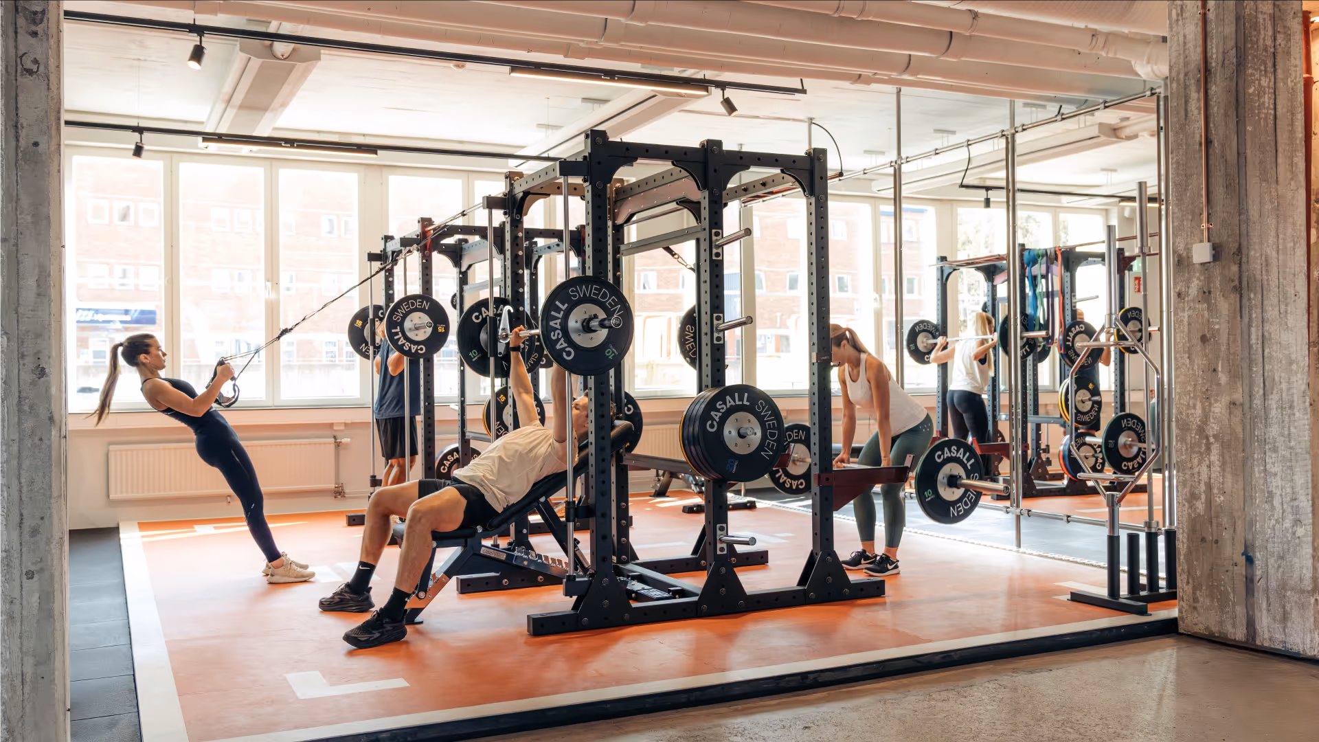 People exercising with barbells and resistance cables in a modern gym with large windows and mirrors.