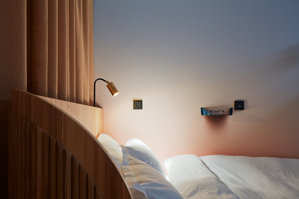 Modern bedroom corner with wooden headboard, white pillows and bedding, a small reading lamp, and a pair of glasses on a wall-mounted shelf.