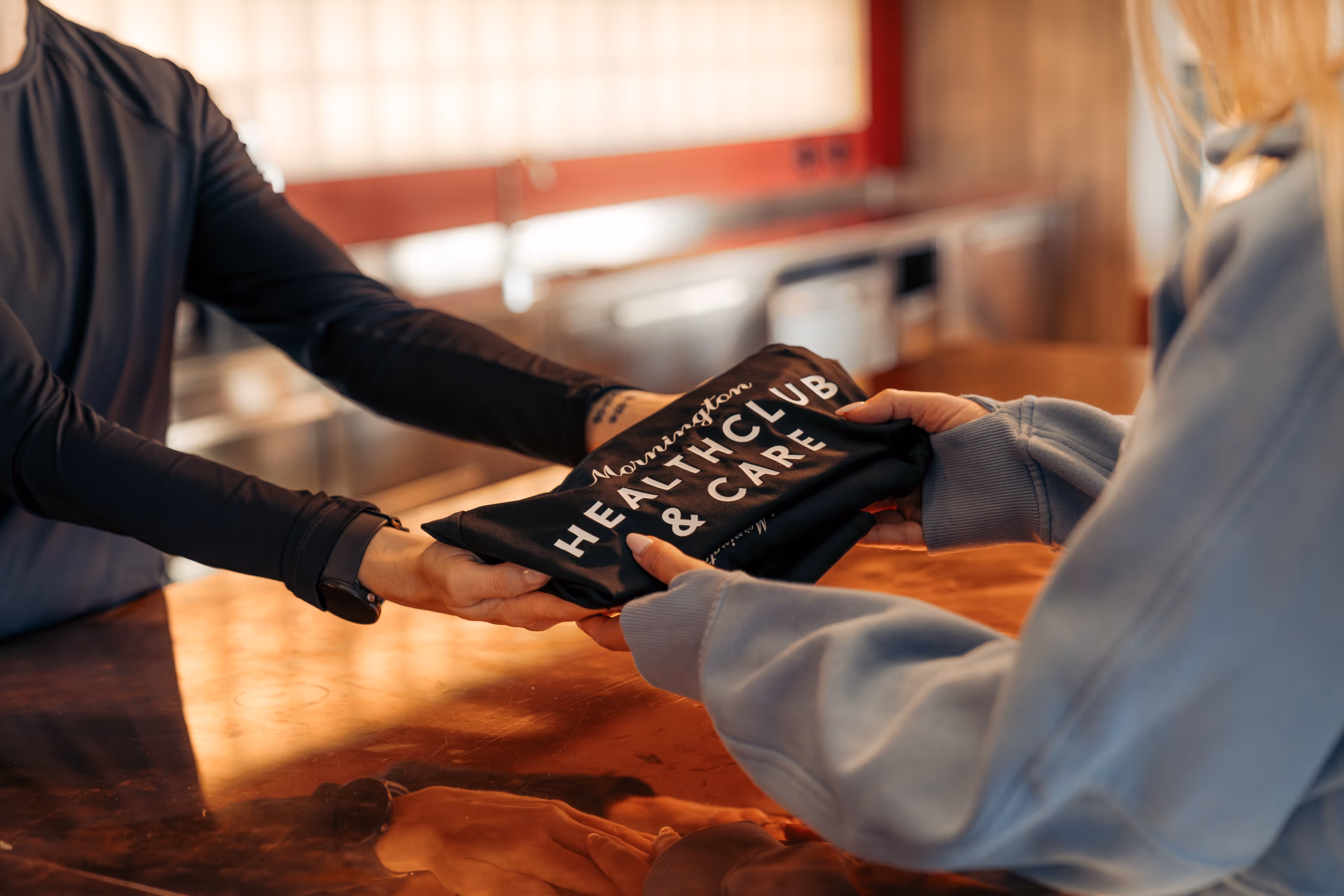 Person in dark long sleeves handing a folded shirt with 'Mornington Health Club & Care' text to another person in a light-colored sweatshirt across a wooden counter.