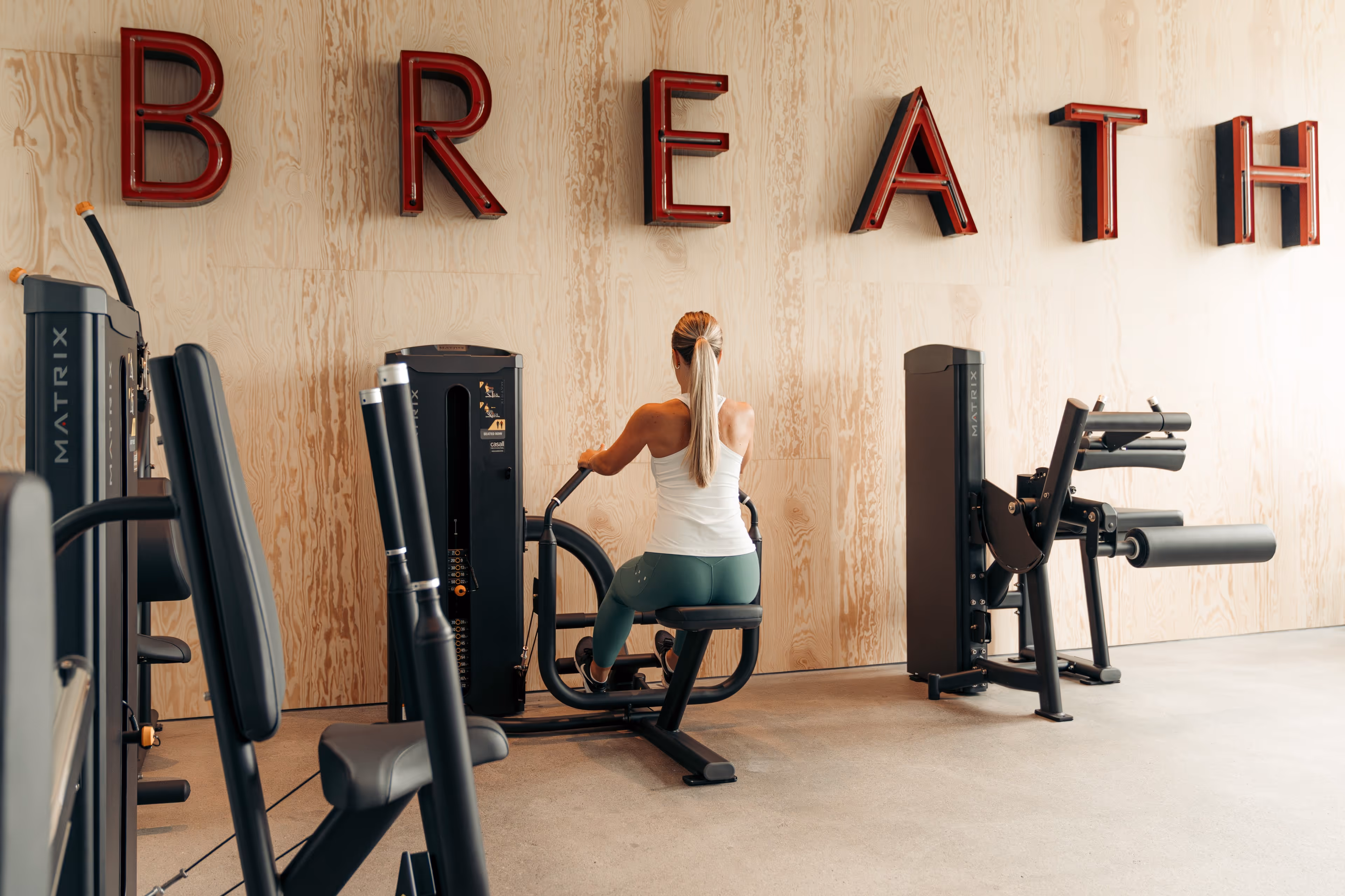 Woman with long blonde hair using a seated rowing machine in a gym with wooden wall and large red letters spelling BREATH.