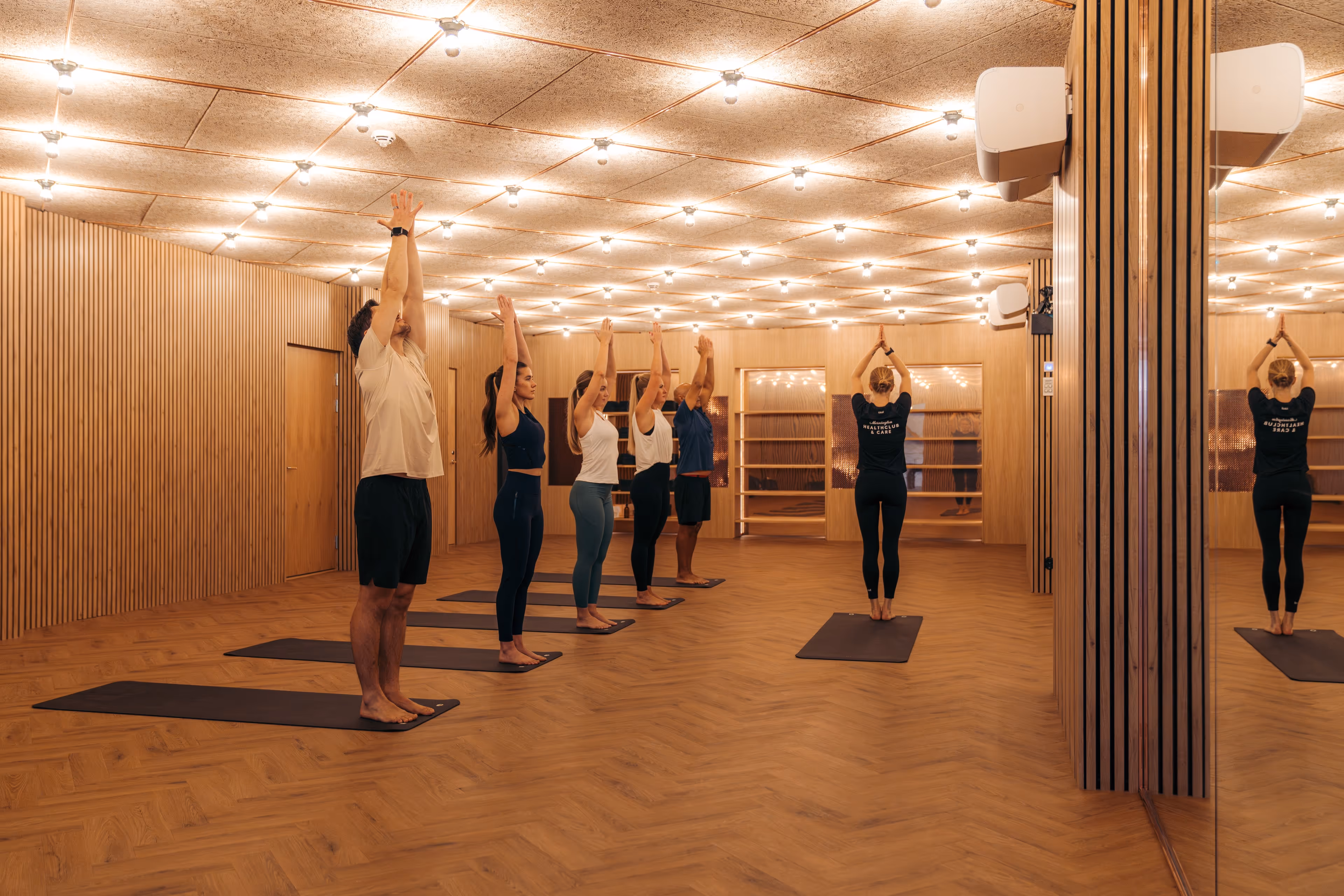 Group of six people in a yoga class standing with arms raised in a modern, warmly lit studio with wooden walls and floor.