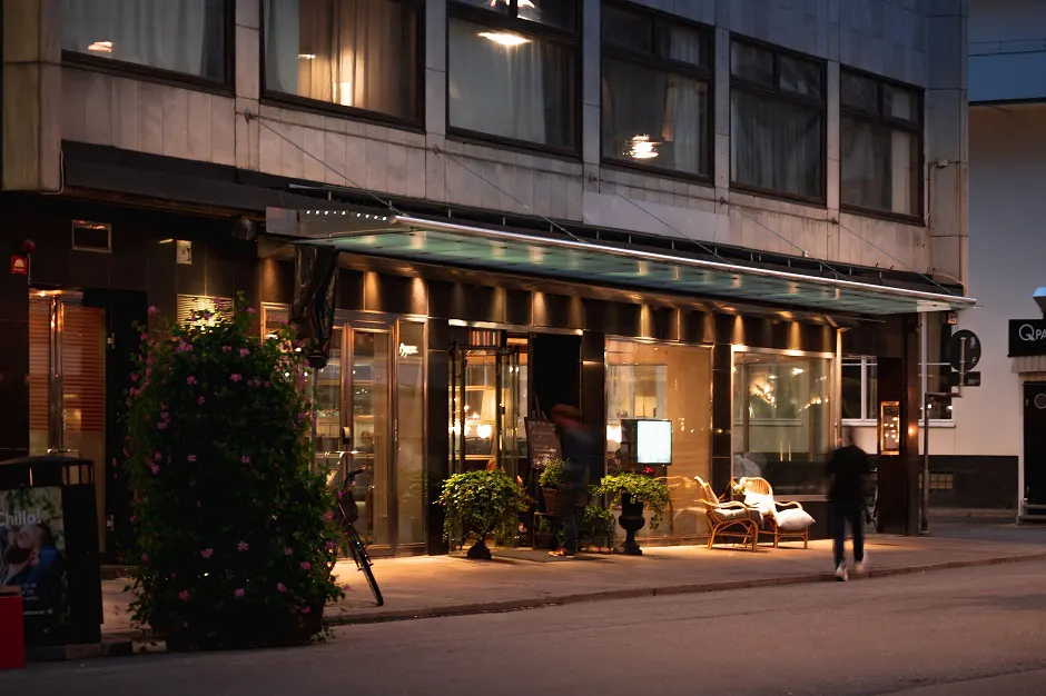 Evening scene of a warmly lit restaurant entrance with outdoor seating and potted plants on a city street.