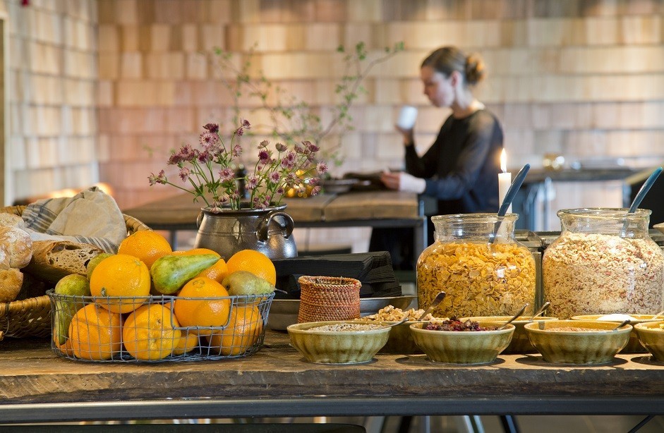 Breakfast table with bowls of cereals, a basket of oranges and pears, bread, and a woman in the background drinking from a cup.