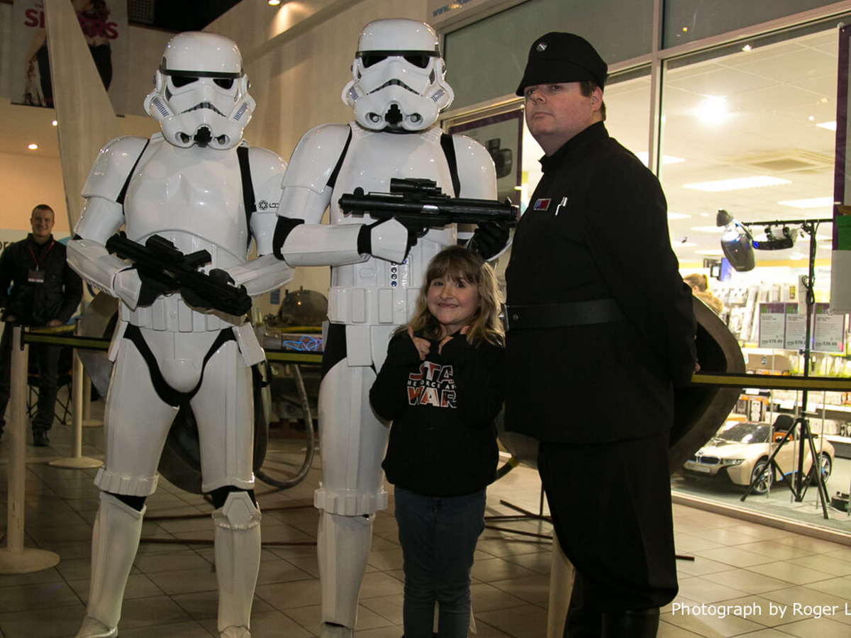 Two people dressed as Star Wars stormtroopers and one person as an Imperial officer posing with a smiling young girl wearing a Star Wars hoodie indoors.