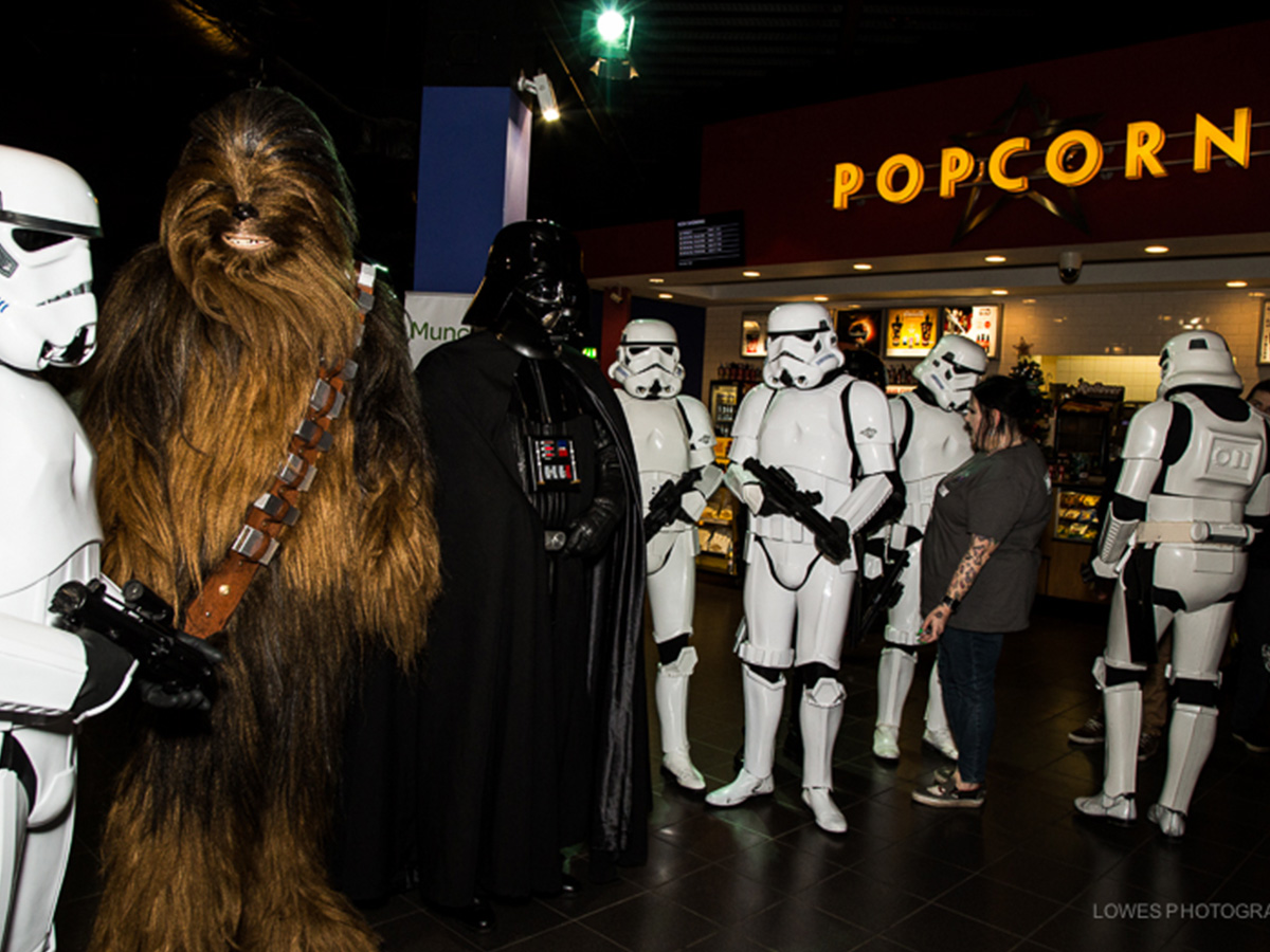 Group of people dressed as Star Wars characters including Chewbacca, Darth Vader, and several stormtroopers inside a cinema near a popcorn stand.