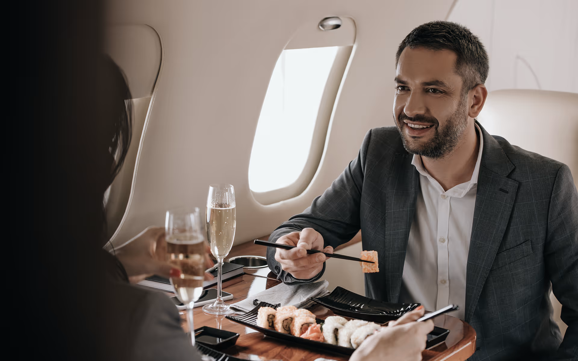 Man in a suit eating sushi with chopsticks and smiling inside a private jet, with a woman holding a glass of champagne across the table.