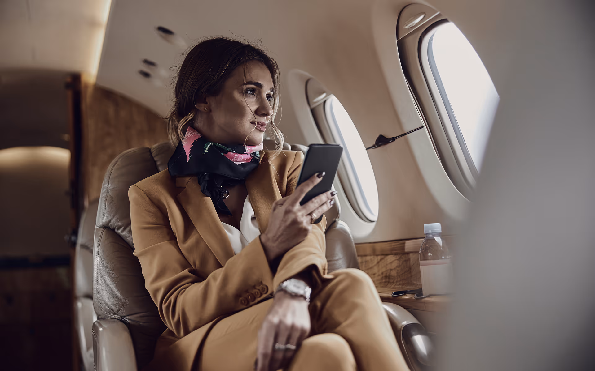 Woman in tan suit sitting in a private jet, holding a smartphone and looking out the window.