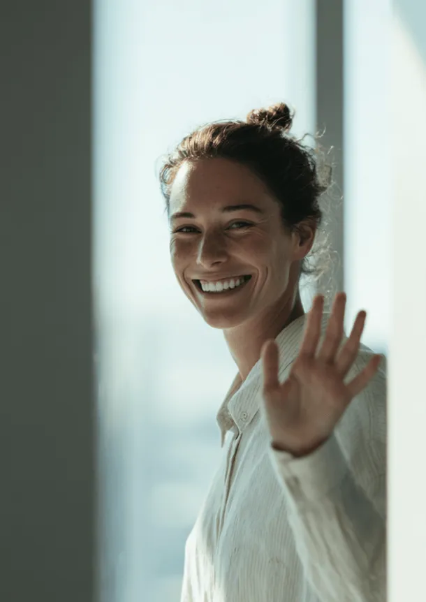A smiling woman waving while standing near a window.