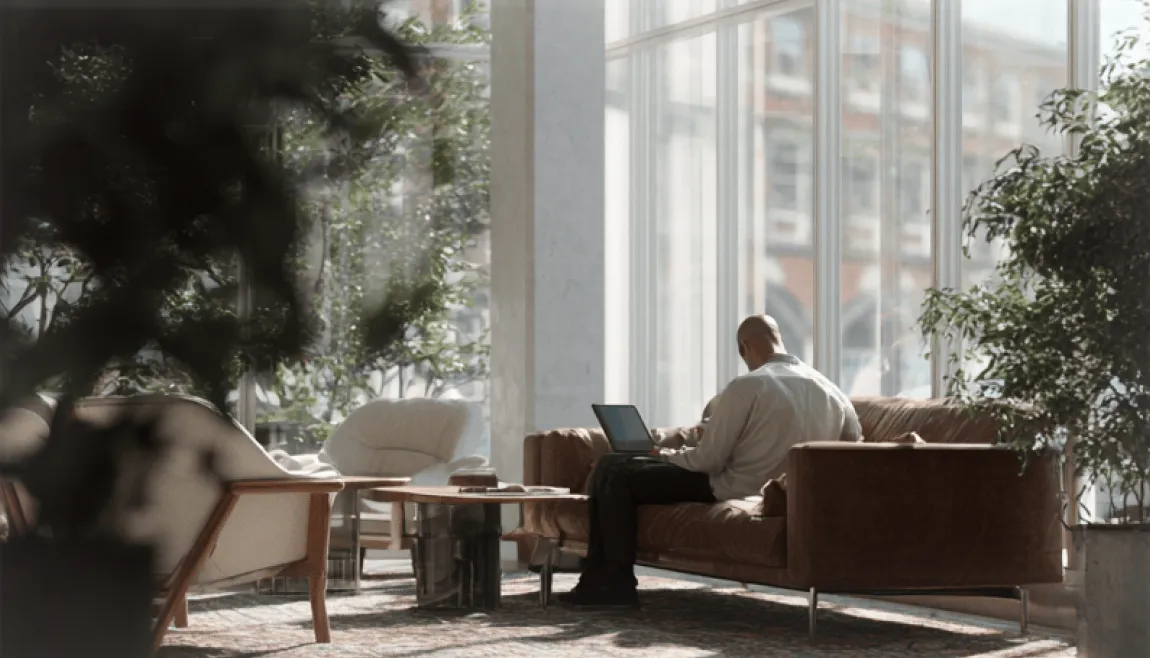 A man using a laptop in a bright lounge with large windows.