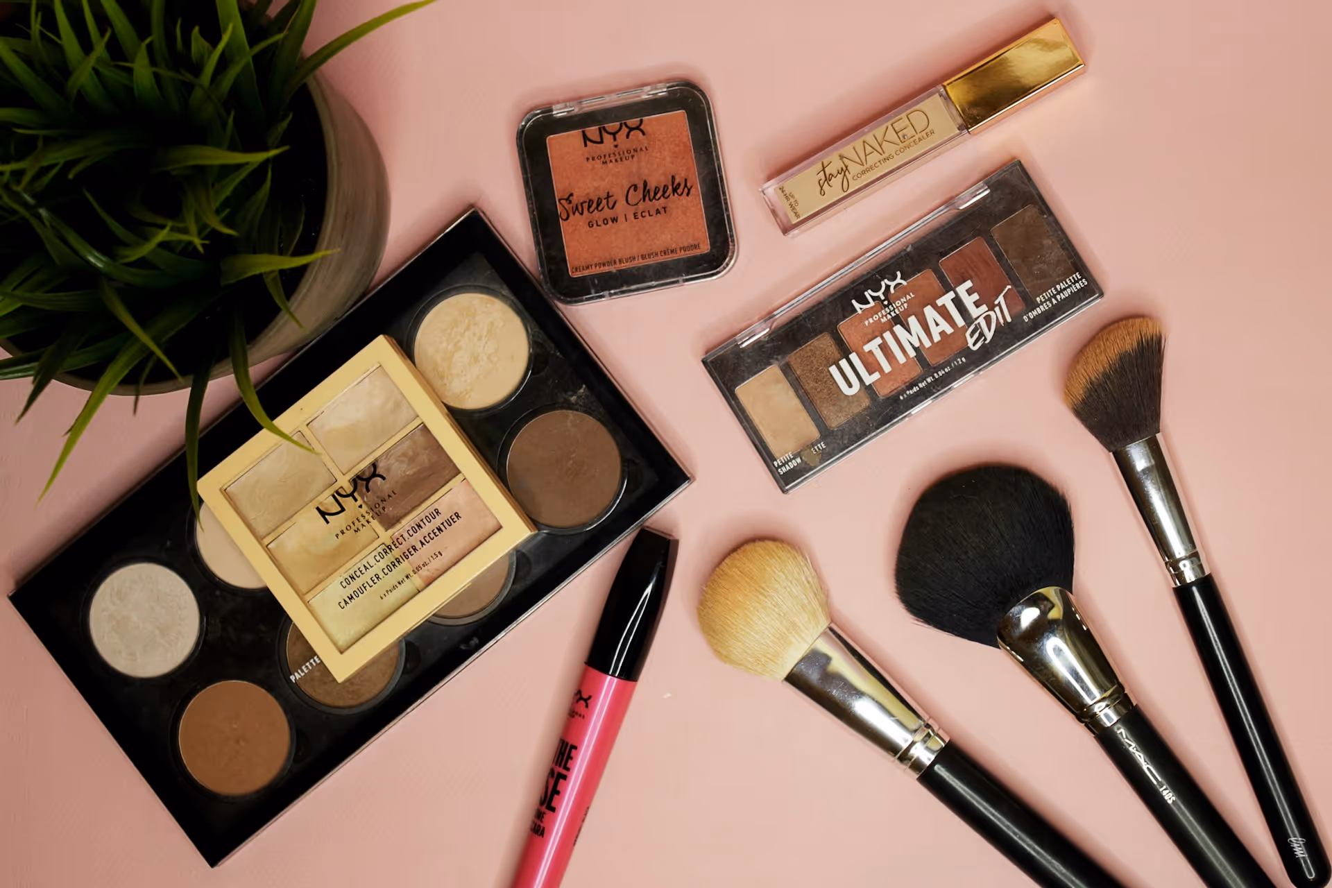 Flat lay of various makeup products including NYX contour palette, blush, concealer, eyeshadow, makeup brushes, and a pink lip gloss on a pink surface with a green plant.