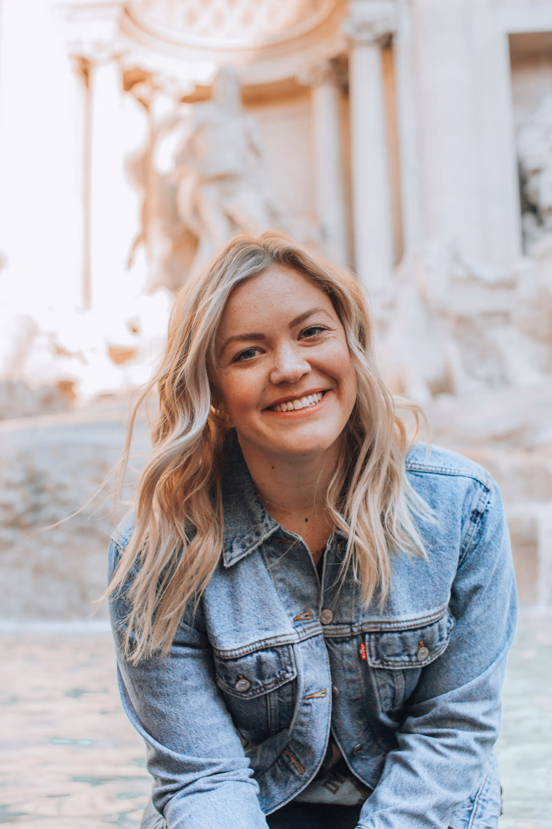 Smiling woman with blonde wavy hair wearing a denim jacket in front of a historic fountain.