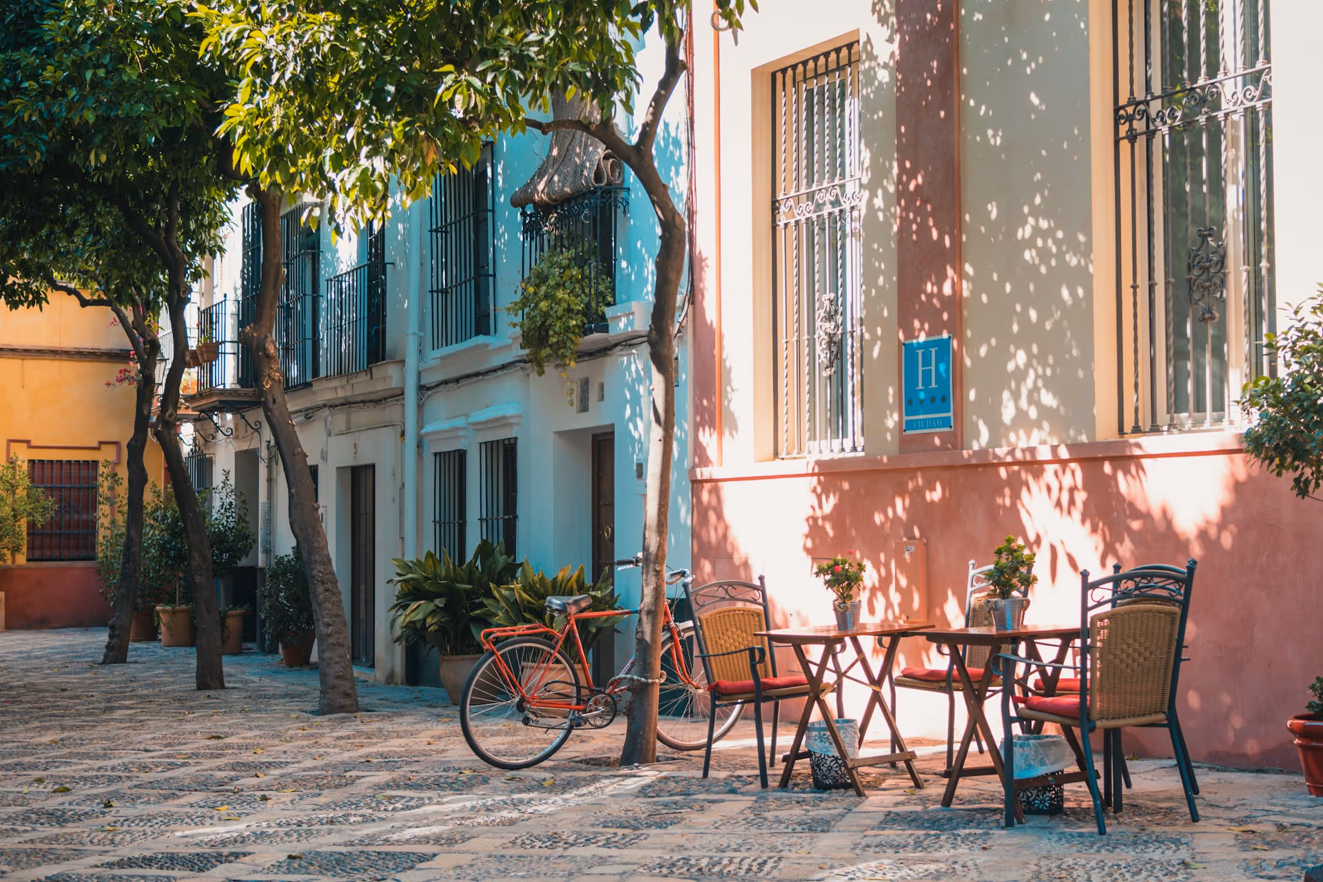 Sunlit outdoor cafe table with four chairs and potted flowers beside a red bicycle under trees in a cobblestone street.