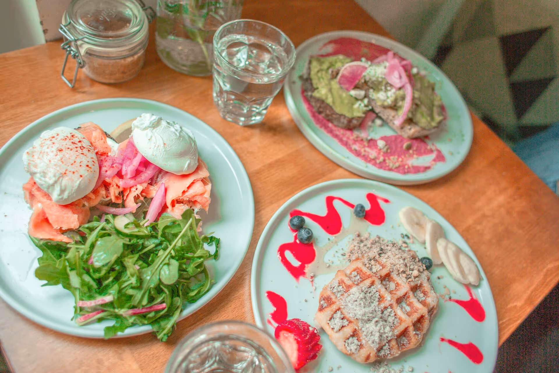 Table with three plates: one with poached eggs, smoked salmon, and salad; one with avocado toast on beet spread; one with waffles topped with nuts, banana slices, blueberries, and strawberry, plus two glasses of water and a jar.